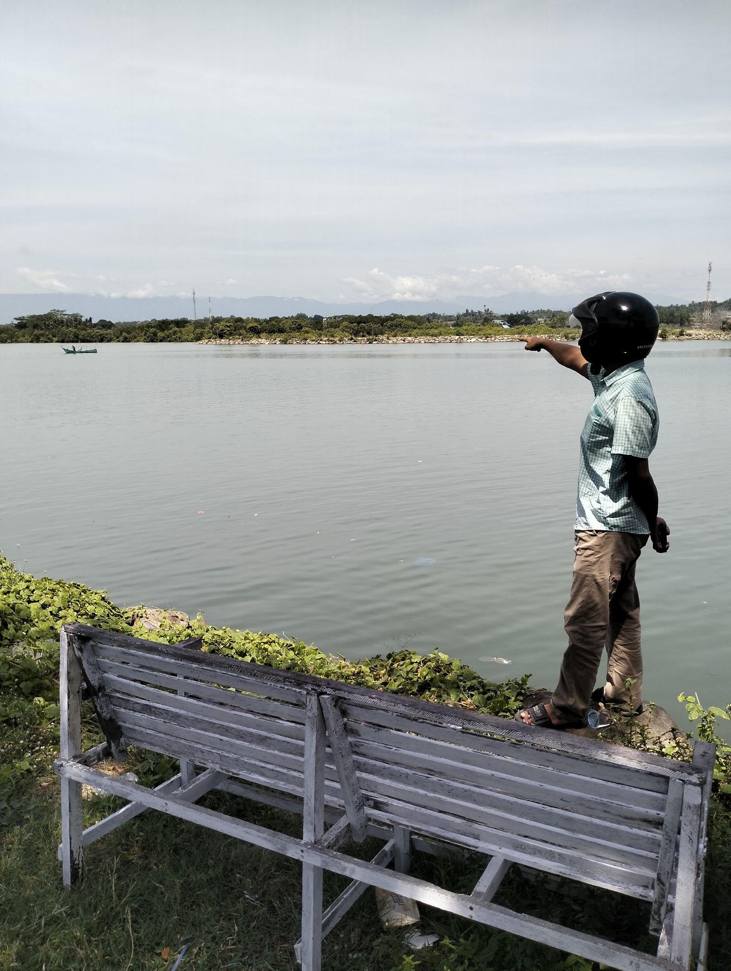 A man wearing a black helmet, a light blue checkered shirt, and khaki pants stands at the edge of a body of water, pointing to the left of the frame. He stands on a grassy bank near a weathered white bench. Debris floats in the water. In the distance, a small boat is visible on the water, with a landmass covered in trees beyond. Further back, mountain ranges are faint in the distance. The location appears to be in Lhokseumawe, Indonesia.
