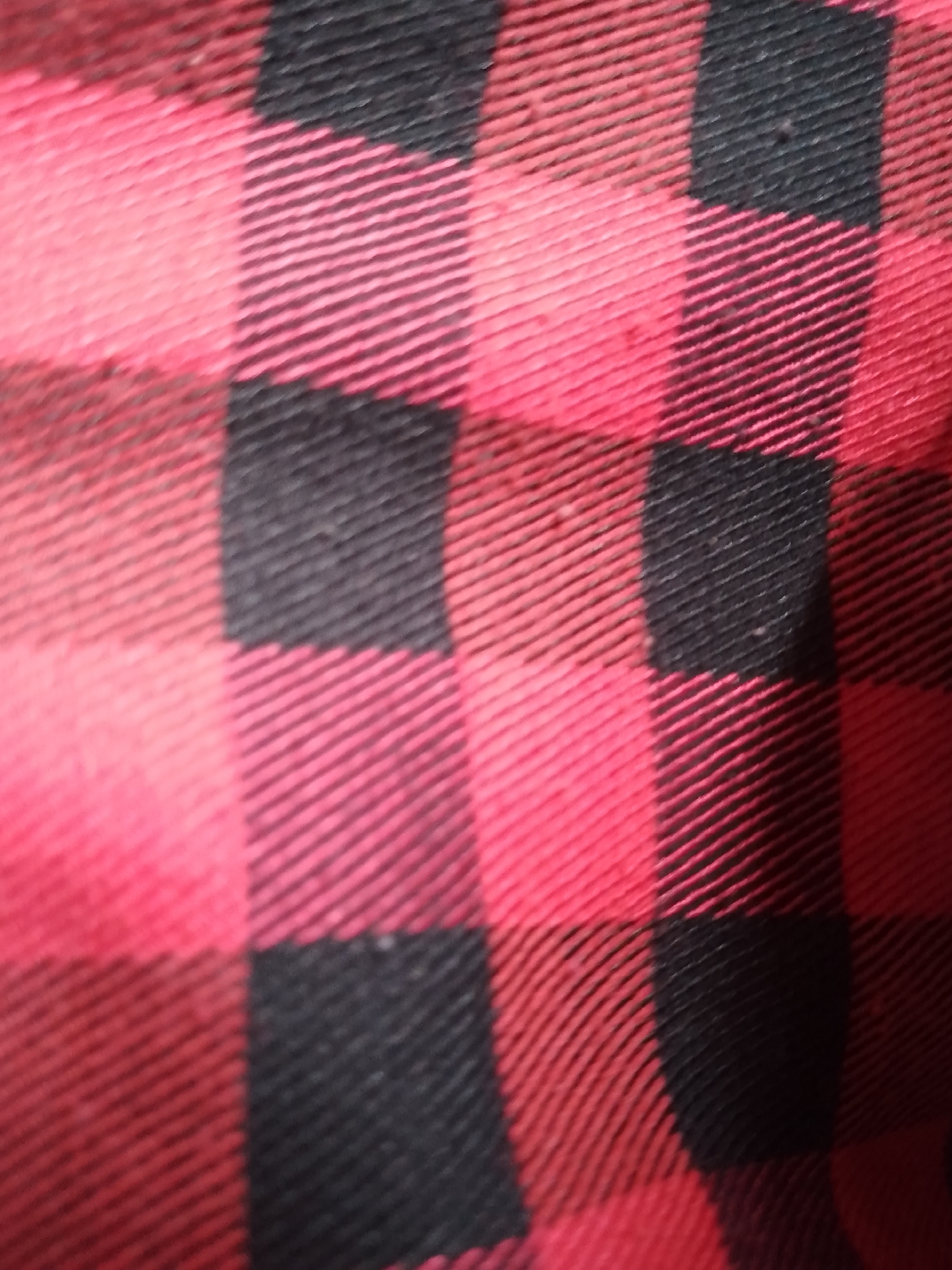 The image shows a close-up of a red and black buffalo plaid fabric. The pattern consists of large squares of solid red alternating with squares of solid black, with thinner intersecting lines of the same colors creating a checkered effect. The fabric appears textured, with visible threads running diagonally. There are no people, objects, actions, interactions, or environmental details visible in the image. The image does not depict any location in Santa Ana, Venezuela.