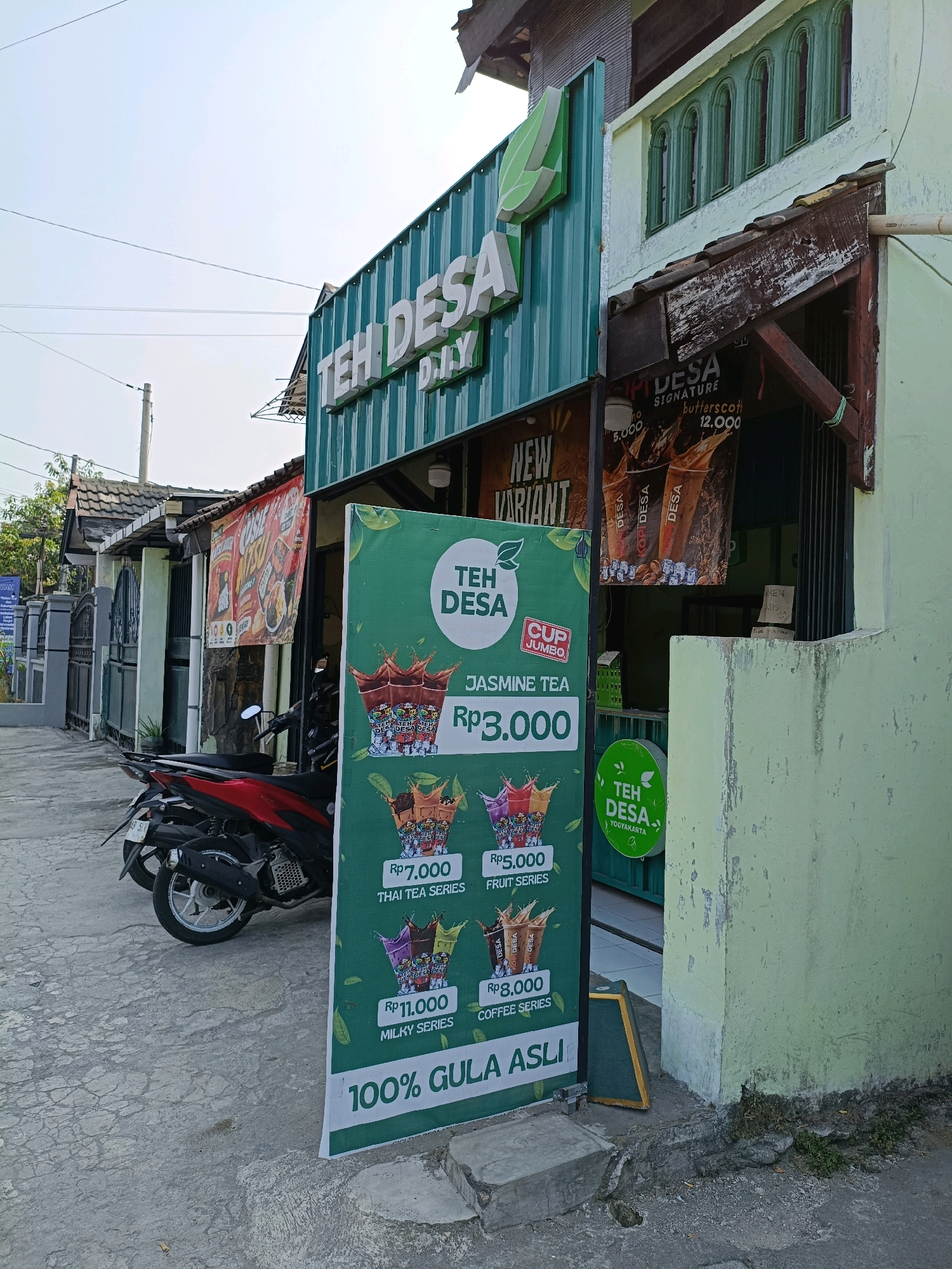 The image captures a street-level view of a business called "TEH DESA" in Maguwoharjo, Indonesia. The storefront is characterized by a green corrugated metal facade with prominent white lettering that reads "TEH DESA D.I.Y." A large green vertical sign displays a menu of tea options, including "JASMINE TEA Rp3.000," "FRUIT SERIES Rp5.000," "COFFEE SERIES Rp3.000," "THAI TEA SERIES Rp7.000," and "MILKY SERIES Rp11.000." The sign also prominently features the slogan "100% GULA ASLI." To the right of the entrance, a circular sign with green text states "TEH DESA YOGYAKARTA." A red motorcycle is parked in front of the establishment. In the background, other shops and buildings line the street, with a red banner featuring Indonesian text visible on one of them. The scene appears to be an outdoor commercial area on a bright day.