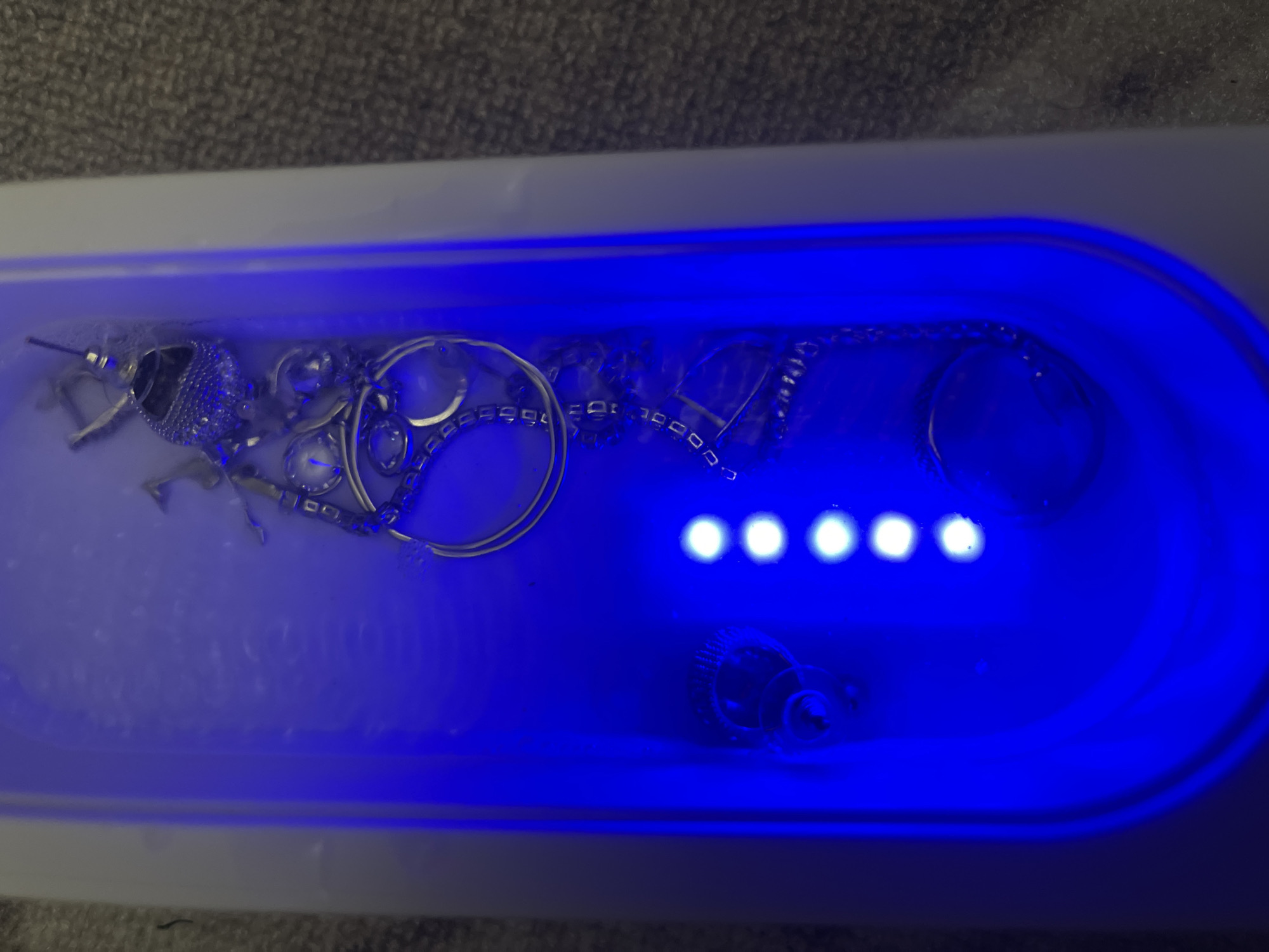 The image shows a white, rectangular ultrasonic cleaner filled with water and various pieces of jewelry, including necklaces, rings, and earrings. Five bright white LED lights illuminate the water from below. The background is a blurry gray surface. The image does not show any identifiable information relating to the city of Chaikovsky, Russia.
