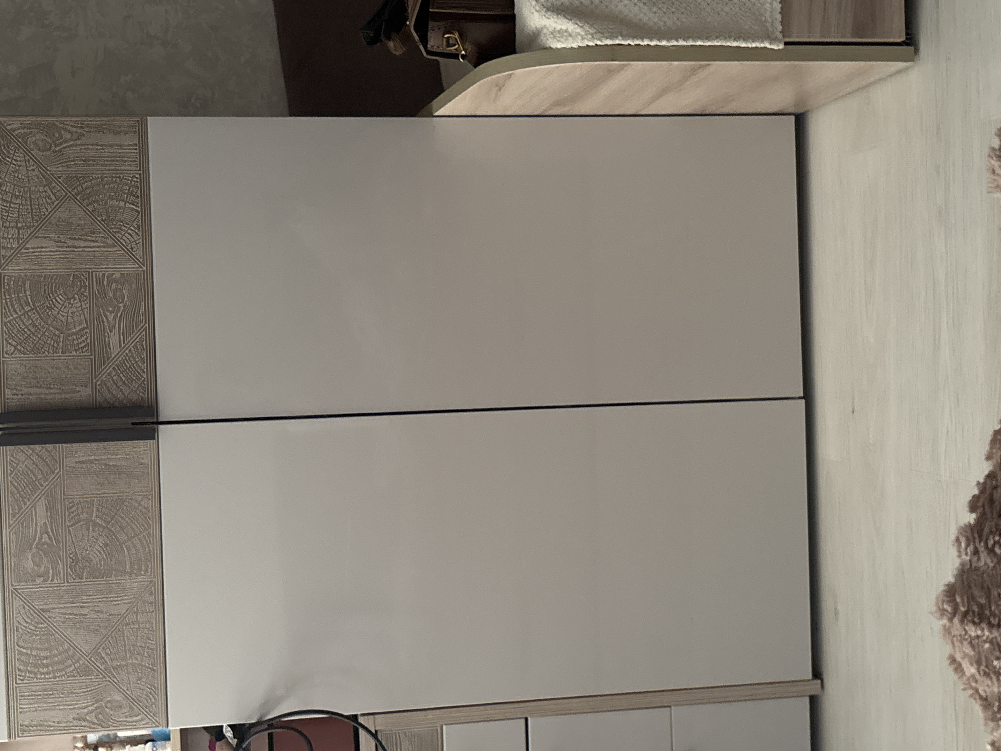 The image displays a close-up view of a modern wardrobe, possibly located in a bedroom. The wardrobe features two large, smooth, light-colored doors that dominate the center of the frame. Above the doors, there is a decorative panel with a wood-grain pattern in shades of beige and brown, arranged in a geometric design. A dark vertical line, likely a seam or handle, divides the two wardrobe doors. To the left of the wardrobe, partially visible, are drawers or compartments with a light wood trim. To the right, a portion of a bed or nightstand is visible, featuring a light-colored textured fabric and a small brown leather bag with a brass clasp. The floor appears to be light-colored laminate or wood. The background wall is a neutral, textured gray. The overall impression is one of a clean, contemporary living space. The scene does not provide any specific indicators to pinpoint its location in Vsevolozhsk, Russia, or any other city.