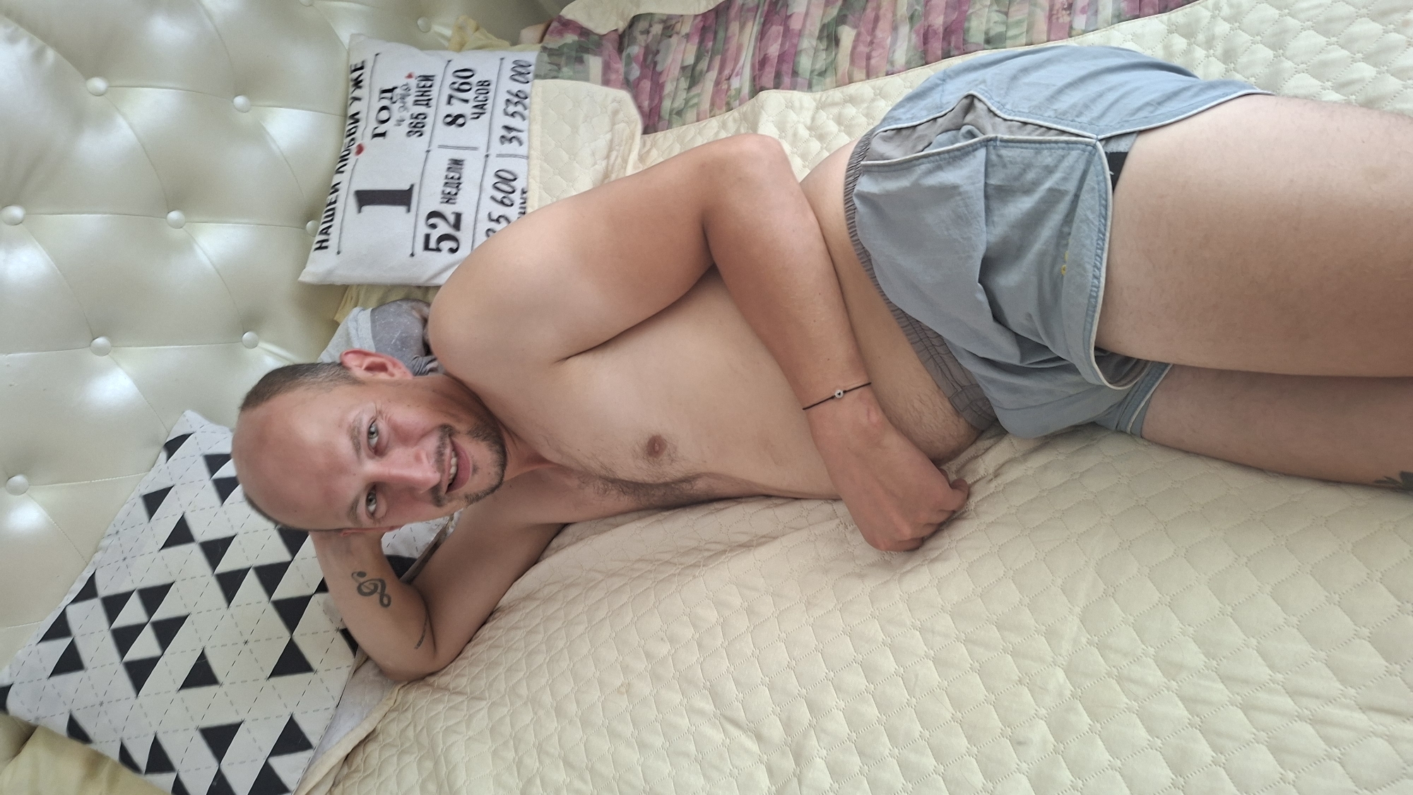 A shirtless man with a shaved head and a beard lies on a bed, propped up by pillows. He is wearing light blue shorts. His left arm is behind his head, and his right arm rests on his stomach. A thin black bracelet with a small white charm is on his right wrist. He is smiling and looking at the camera. Behind him, a white tufted headboard forms the backdrop. To his left, a patterned pillow with black and white triangles is visible. To his right, another pillow with text and numbers is placed against the headboard. The bed is covered with a quilted, light-colored comforter. The overall scene appears to be an indoor bedroom setting. No specific location within Myadzyel, Belarus, can be determined from the image.