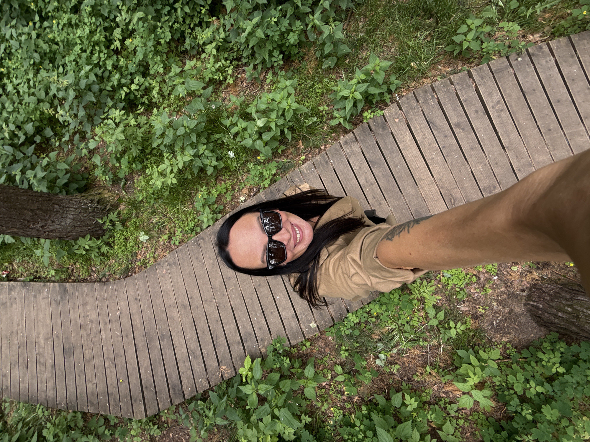 A high-angle, full-body selfie of a woman with long dark hair, wearing sunglasses and a tan jacket, is taken while she is standing on a wooden boardwalk. The boardwalk curves to the left and is surrounded by lush green foliage and trees. The scene appears to be an outdoor natural setting, possibly a park or trail within Chaikovsky, Russia, given the context. The woman is smiling directly at the camera, and her arm, holding the phone, extends into the lower part of the frame, revealing a tattoo on her forearm.