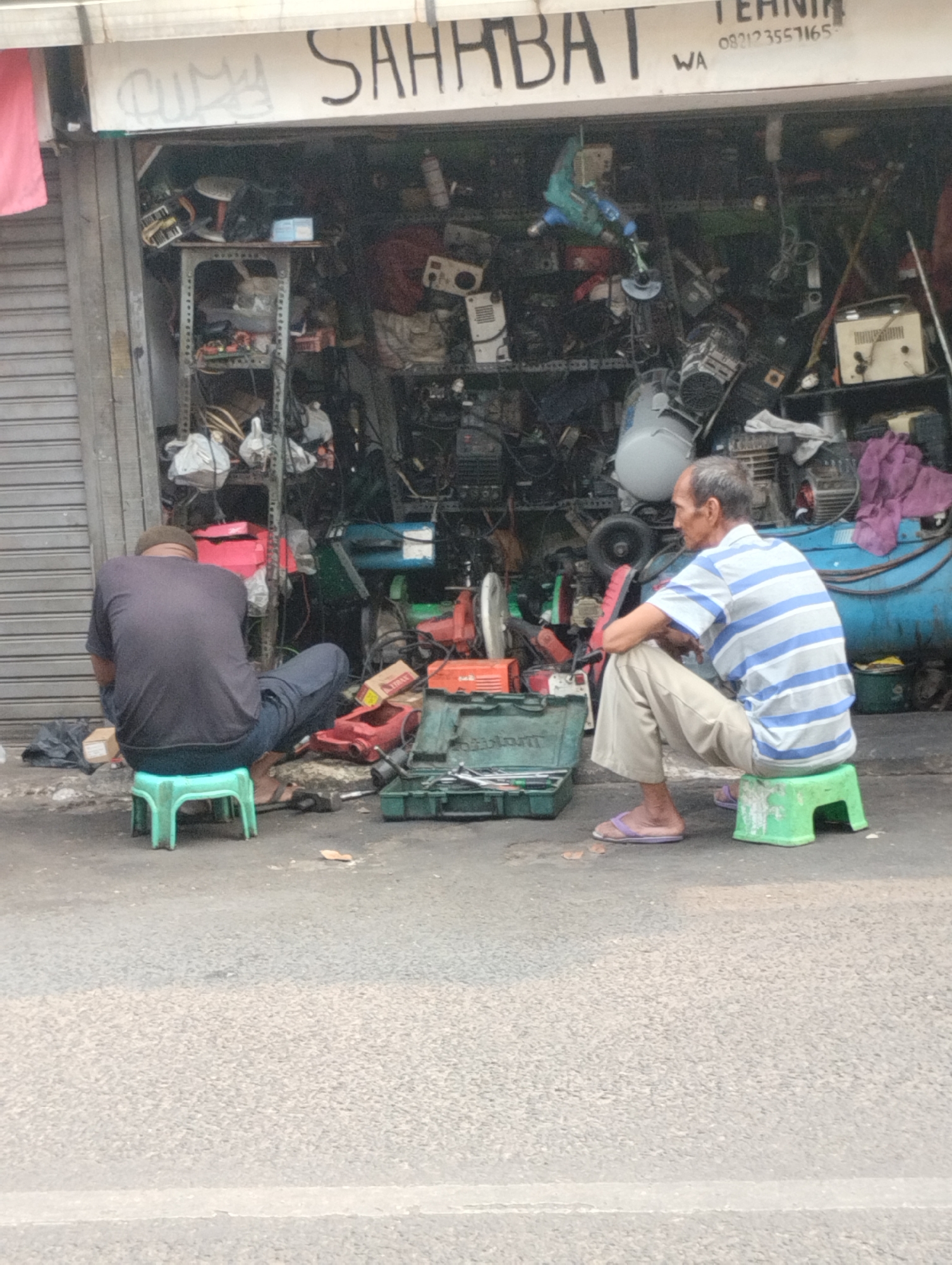 Two men are sitting on green plastic stools in front of a shop in Bekasi, Indonesia. The shop is filled with various mechanical parts and equipment. The man on the left is wearing a dark t-shirt and pants and is facing away from the camera, looking towards the shop. The man on the right is wearing a striped t-shirt and light-colored pants and is facing left, looking towards the shop. A toolbox is open on the ground in front of him, containing various tools. The shop has a sign above the entrance that reads "SAHABAT TEKNIK" and a phone number. The overall scene suggests a repair shop or a seller of used mechanical parts.