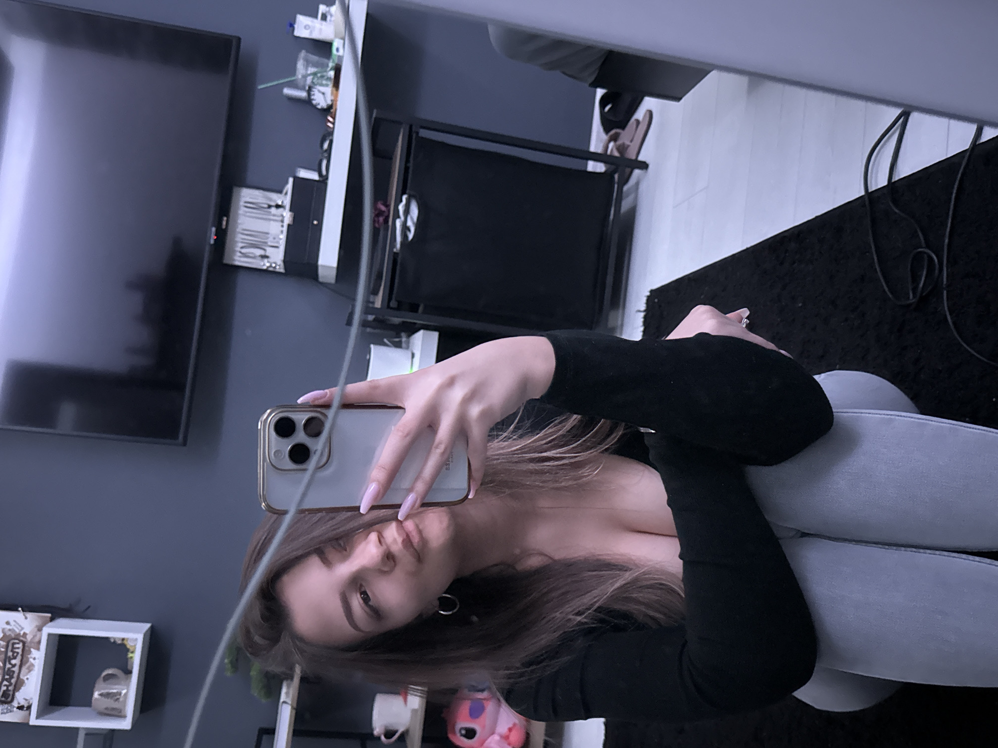 A medium shot depicts a fair-skinned person with brown hair, dressed in a black long-sleeved shirt and grey leggings, taking a selfie in a mirror. The phone being used is a gold-trimmed iPhone with three rear cameras. The person's legs are crossed, and the room includes a black television mounted on a grey wall, white floating shelves with decorations, a black wireframe storage unit, and a black rug on light-colored wood flooring. The location is possibly in Sofia, Bulgaria, but cannot be determined without more information.
