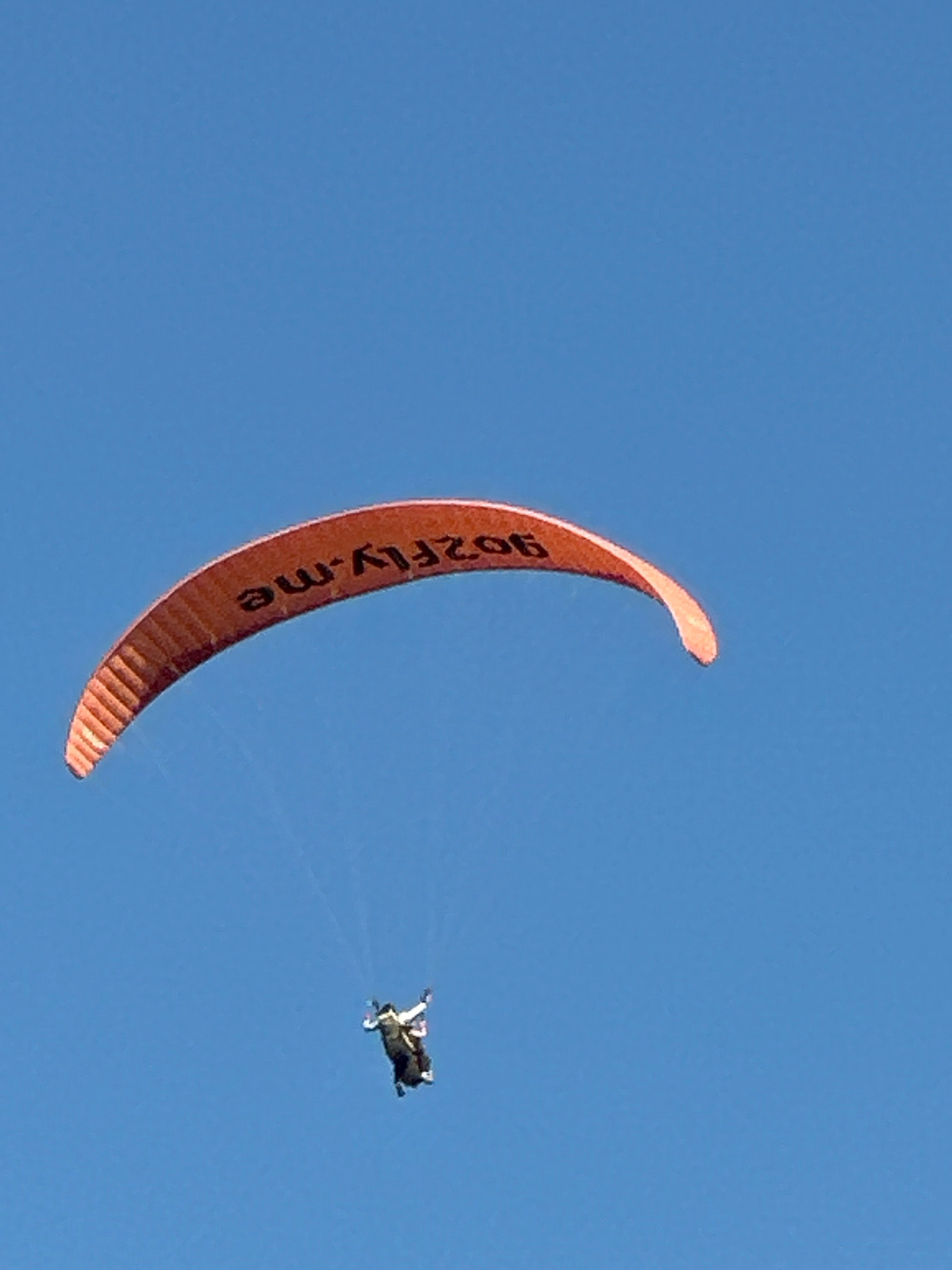 An orange paraglider with the text "902FLY.ME" written on it is seen against a clear blue sky. Below the paraglider, a person is suspended, appearing to be in freefall. The image is taken from below, looking up at the paraglider and the individual. The location is Boreti, Montenegro.
