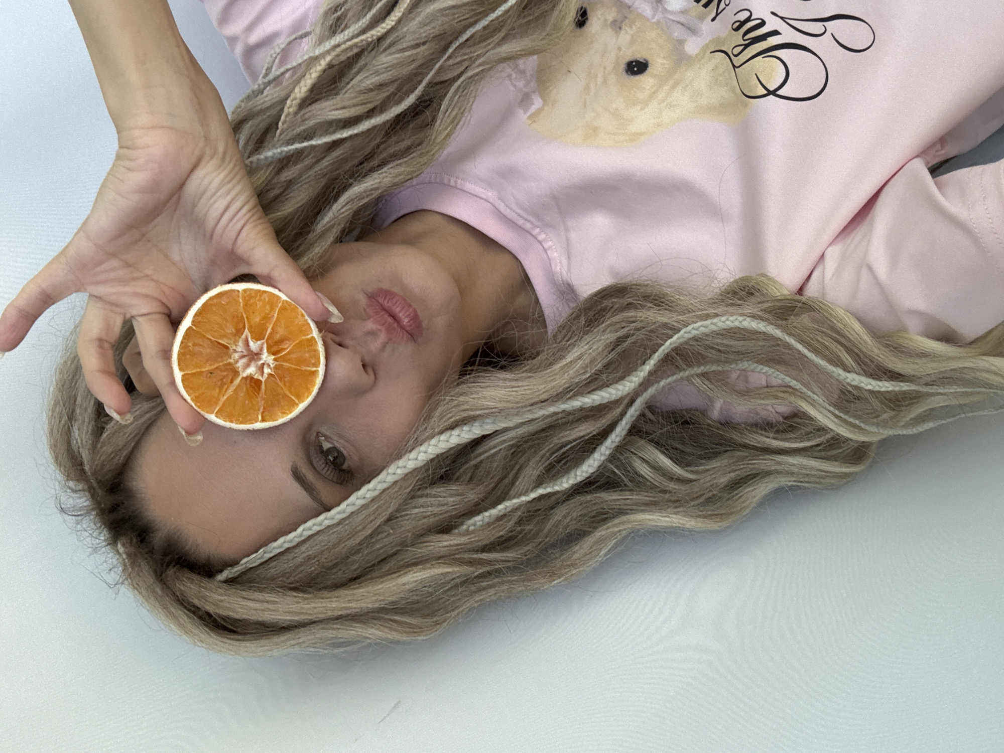 A woman lies on a white surface, facing upwards. She is wearing a light pink shirt with a teddy bear graphic and text on it. The woman has long, blonde hair with white braids incorporated. In her right hand, she holds a dried orange slice in front of her right eye. She is facing the camera with her lips pursed. The location is Unknown, Unknown.