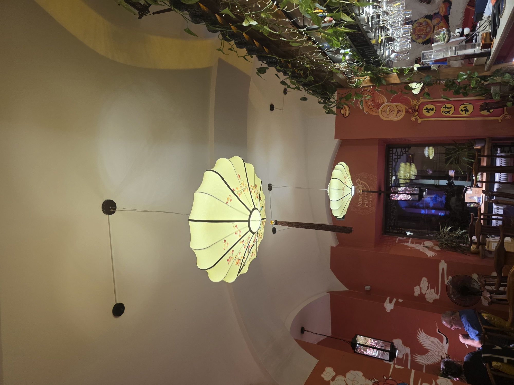 The image depicts the interior of an East Asian-themed restaurant, located in the city of Unknown, Unknown. The scene is illuminated by ambient lighting, suggesting an evening setting.
The ceiling, predominantly off-white with arched sections, features several pendant light fixtures. Two large, pale yellow, scalloped lanterns with dark frames and red/orange floral patterns hang centrally from the ceiling; the larger one includes a dark brown decorative tassel. A smaller, similar lantern is visible further back. Several smaller black-framed rectangular lanterns, some appearing to contain illuminated cherry blossom designs, are also suspended from the ceiling and mounted on walls.
The walls are painted a reddish-orange hue and adorned with large white murals depicting cranes and traditional cloud patterns. A distinct section of the wall features a vertical mural of gold-colored fruit and circular coins, with a prominent red and gold scroll displaying the Chinese characters "生財利" (shēng cái lì), meaning "generate wealth and profit." A sign on the wall clearly displays "KUNG FU RAMEN" in English and "功夫拉面" in Chinese characters.
In the lower-left quadrant, two individuals are seated at a dark wooden table. One person, seen from the back, has dark hair. Adjacent to them, a man with light, short hair and wearing a blue shirt is visible from the side, holding chopsticks. A black standing fan is positioned on the floor nearby.
To the right, a bar area is present, featuring numerous clear wine or cocktail glasses hanging inverted from a rack. Green vining plants, possibly pothos, are draped along the top of the bar structure, interwoven with string lights. A small panda figurine is visible on the bar counter. Dark wooden chairs and tables are scattered throughout the visible dining area.