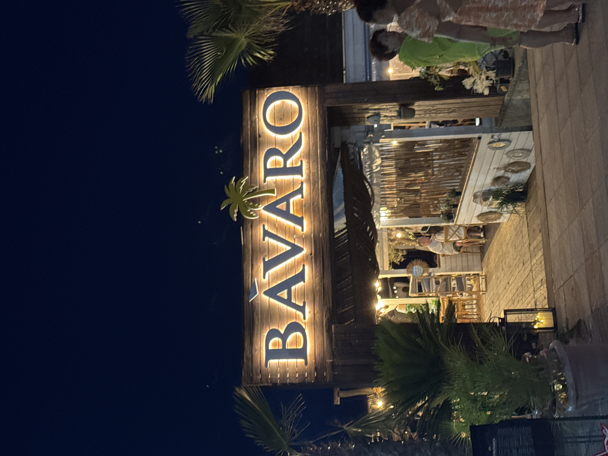 The image shows the exterior of a restaurant at night, with the illuminated sign "BÀVARO" prominently displayed above the entrance. The sign is mounted on a wooden facade decorated with a stylized palm tree. The restaurant appears to be open, with tables and chairs visible inside and on a wooden deck. Several people are seen in and around the restaurant. Two women are standing in the foreground, facing away from the camera. A man is seated at a table inside. The area is surrounded by palm trees and other greenery, with decorative lighting creating a warm ambiance. The scene is set in Burgas, Bulgaria, likely near the coast given the tropical-themed decor and palm trees.
