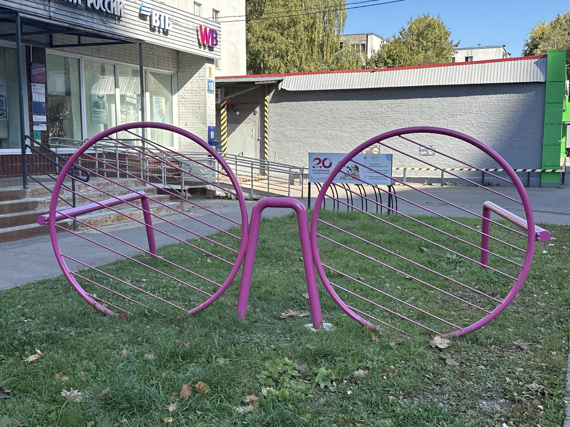 A pink, oversized pair of eyeglasses sculpture stands on a grassy area in front of a building. The building has a facade with horizontal siding and a sign that reads "POST OF RUSSIA". There are windows with merchandise displays visible behind the glass. A ramp with a handrail leads up to the entrance. To the right of the building, there is a grey brick wall with yellow and black striped markings and a closed door. A sign advertising an optician is visible behind the sculpture, with text in Russian and images of people and eyeglasses. The sculpture is made of metal rods forming two large circular frames connected by a bridge-like structure. The ground is covered with green grass and scattered fallen leaves. The sky is blue and clear. The location is identified as Podolsk, Russia.