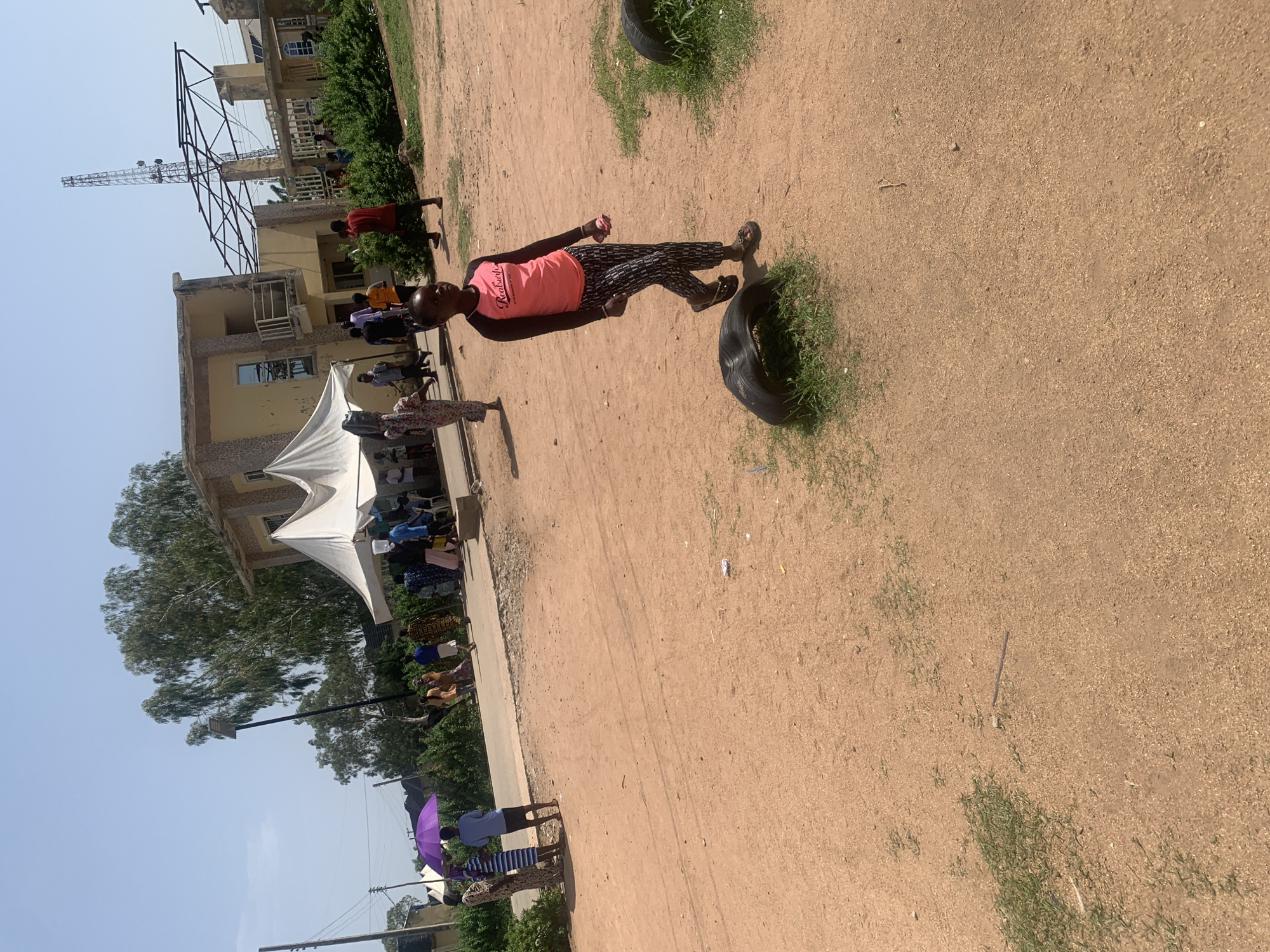The image depicts an outdoor scene in Jalingo, Nigeria, under a clear blue sky. In the foreground, a young girl with dark hair is walking on a dirt path. She is wearing a pink long-sleeved shirt with black sleeves and a graphic print, and patterned black and white trousers. She is also wearing dark sandals. To her left, partially visible on the ground, is a deflated tire with grass growing through it, and another similar tire is visible further to the right.
In the mid-ground, a group of people are gathered near a light yellow, two-story building. A white tent structure is set up in front of the building, and several individuals are visible beneath it, some seated on chairs. A purple umbrella is held by a person standing to the left of the tent. A road runs behind the group of people. The building has balconies and windows and appears to be under construction or renovation, with a large metal framework extending upwards behind it, possibly for a telecommunications tower.
In the background, more buildings and lush green trees are visible. The ground is predominantly a dusty, reddish-brown dirt. The overall impression is a bustling outdoor gathering or event.