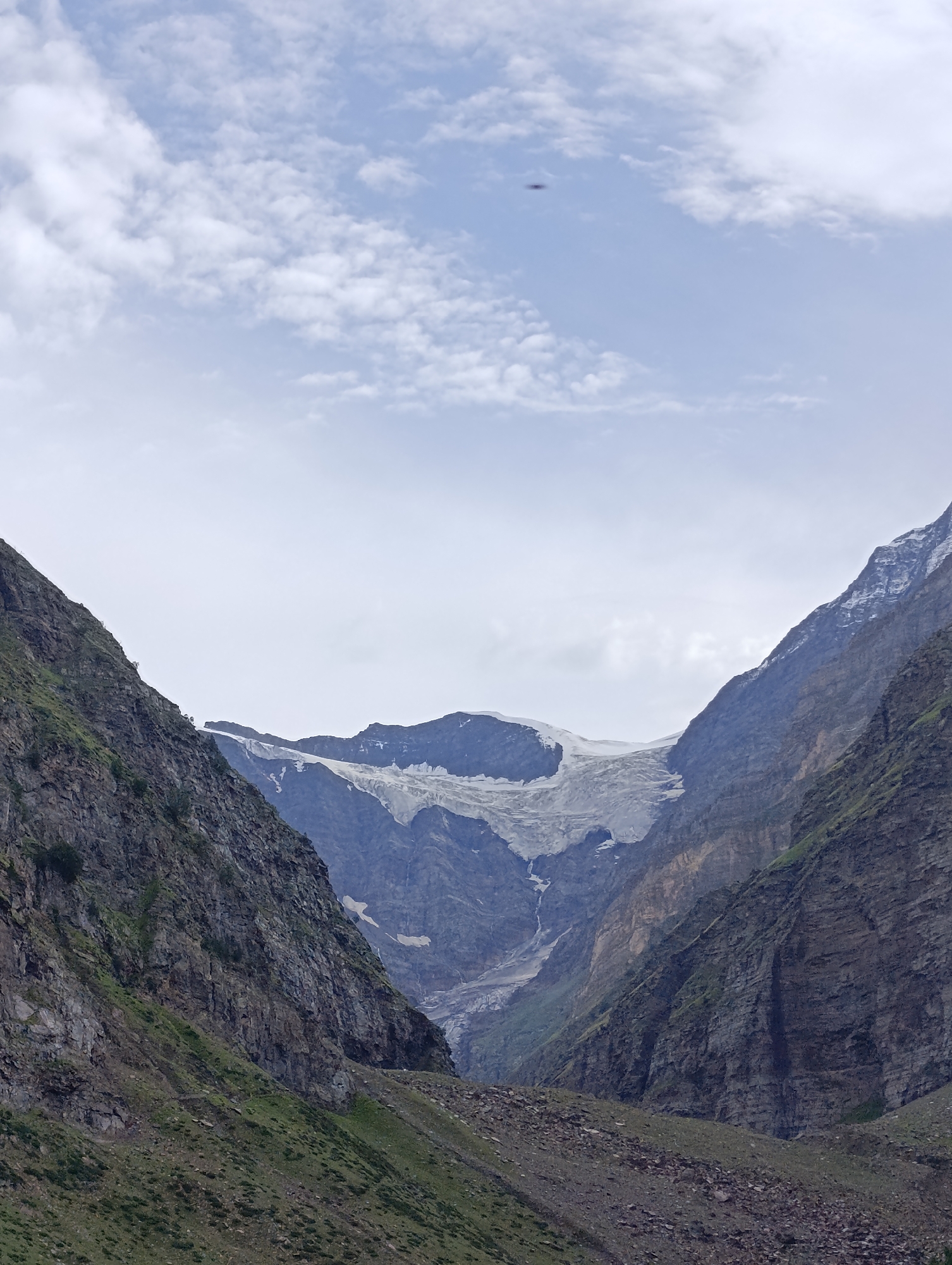 The image shows a panoramic view of a mountain valley in Sissu, India. The foreground features rocky slopes with sparse green vegetation. The midground is dominated by steep, rugged mountains that frame a large glacier at the far end of the valley. The glacier is white and appears to be receding, with a darker, rocky terrain visible beneath and around it. The sky above is partly cloudy with a light blue hue, and a small, dark object, possibly a bird or a drone, is visible in the upper center of the frame. The overall scene conveys a sense of natural grandeur and the raw beauty of a high-altitude landscape.