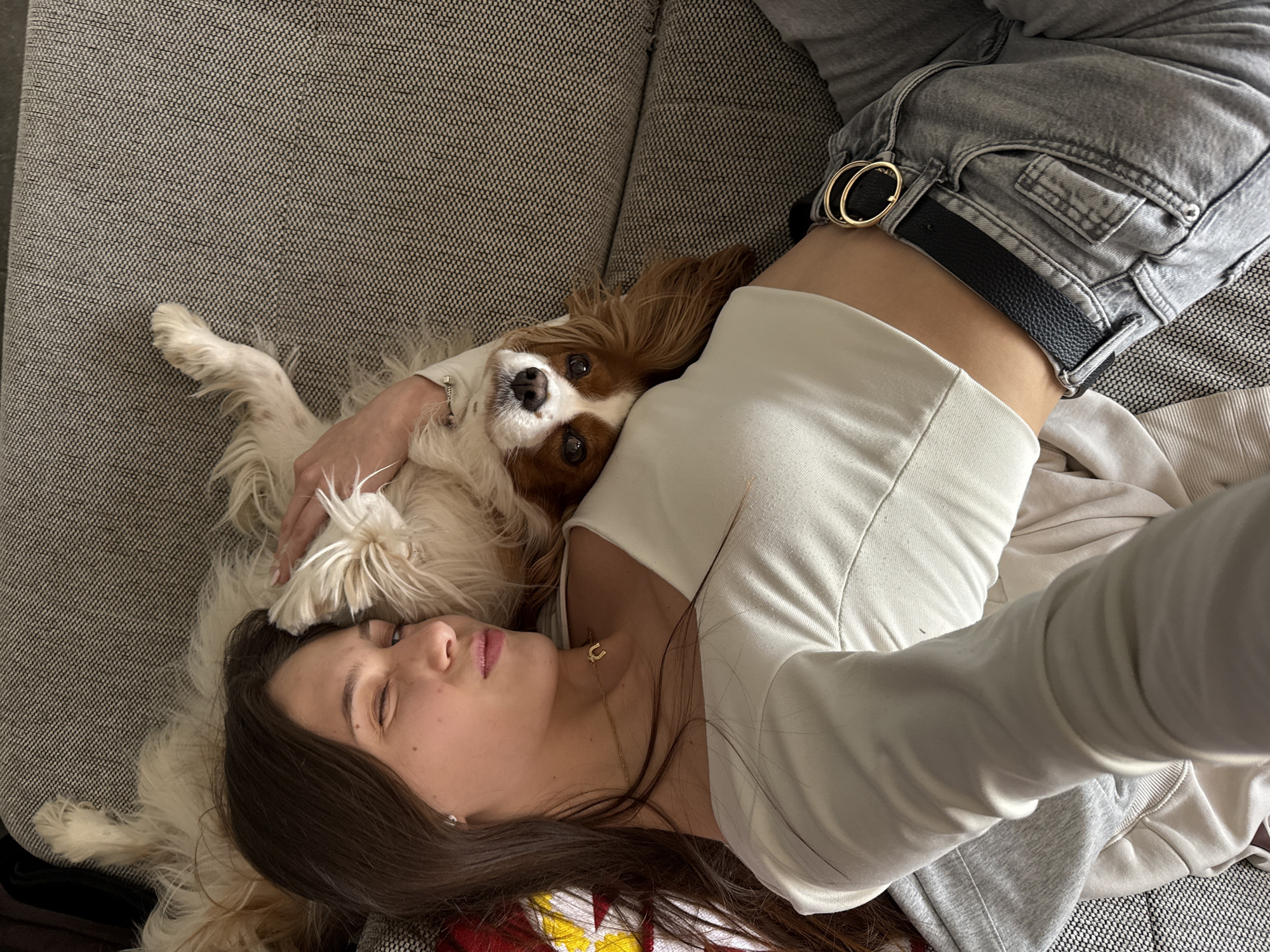 A young woman lies on a gray upholstered sofa with a Cavalier King Charles Spaniel dog. The woman is fair-skinned with long dark hair. She is wearing a white top, gray sweatpants with a black belt and gold buckle, and a gold necklace with a horseshoe pendant. The dog is lying on her chest and head, looking directly at the camera. The woman's left arm is around the dog. Her eyes are closed and she appears to be asleep. The location is unknown, and there is no evidence suggesting the city of Sofia, Bulgaria.
