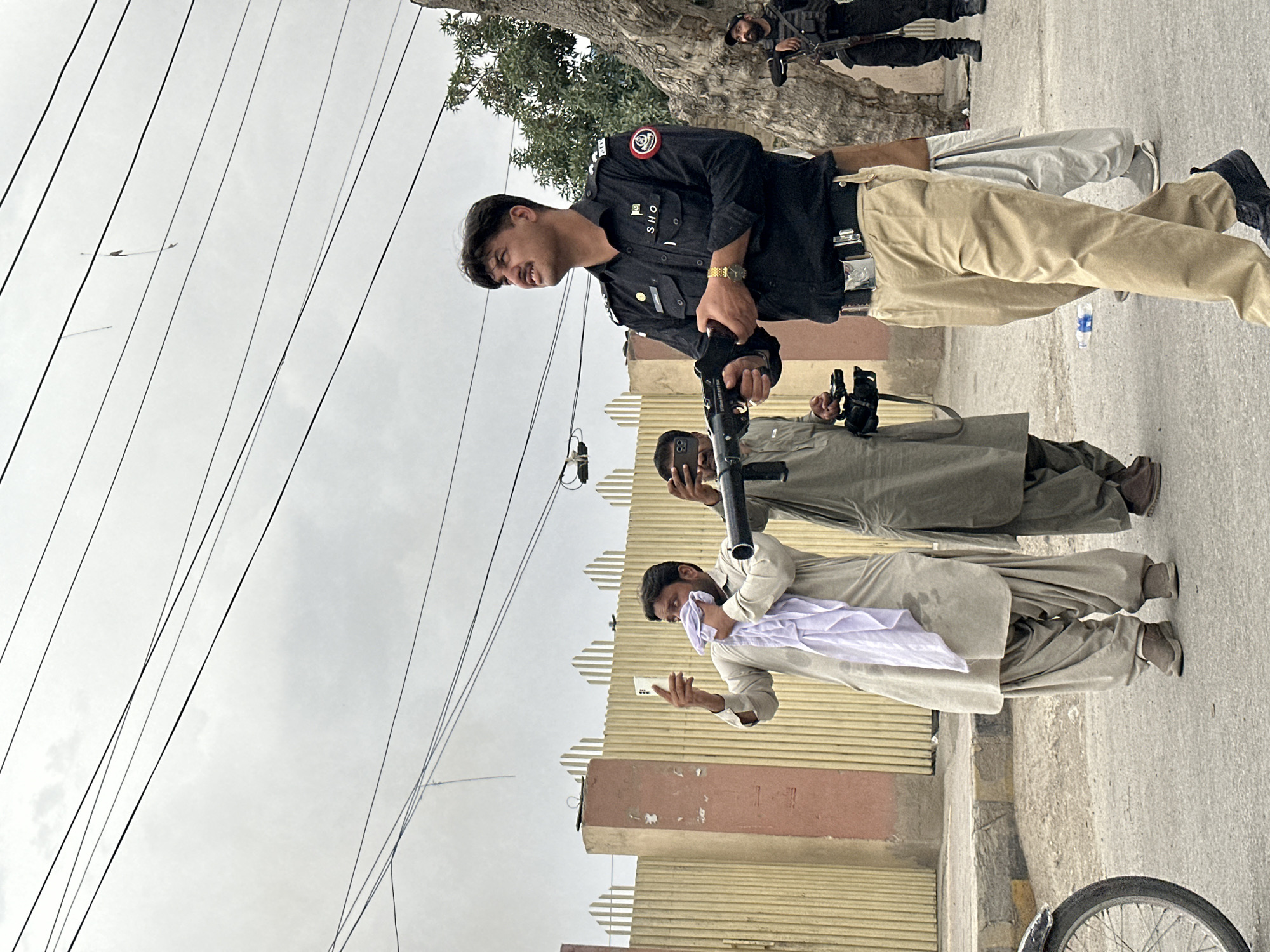 In an urban environment under a cloudy sky, a police officer in a black uniform with "S.H.O" visible on his chest stands holding a rifle. He is wearing tan pants and a watch. To his left, two men are visible; the one in the foreground wears a light-colored traditional garment and holds a white cloth and a mobile phone. The man behind him wears a darker traditional garment and holds a camera. In the background, a tan fence with vertical slats runs horizontally across the image, with a concrete pillar to the left. Further in the background, another uniformed officer with a rifle stands near a tree trunk. The ground is paved, and a bicycle wheel is partially visible on the left side of the frame. A plastic water bottle is on the ground in the mid-ground.