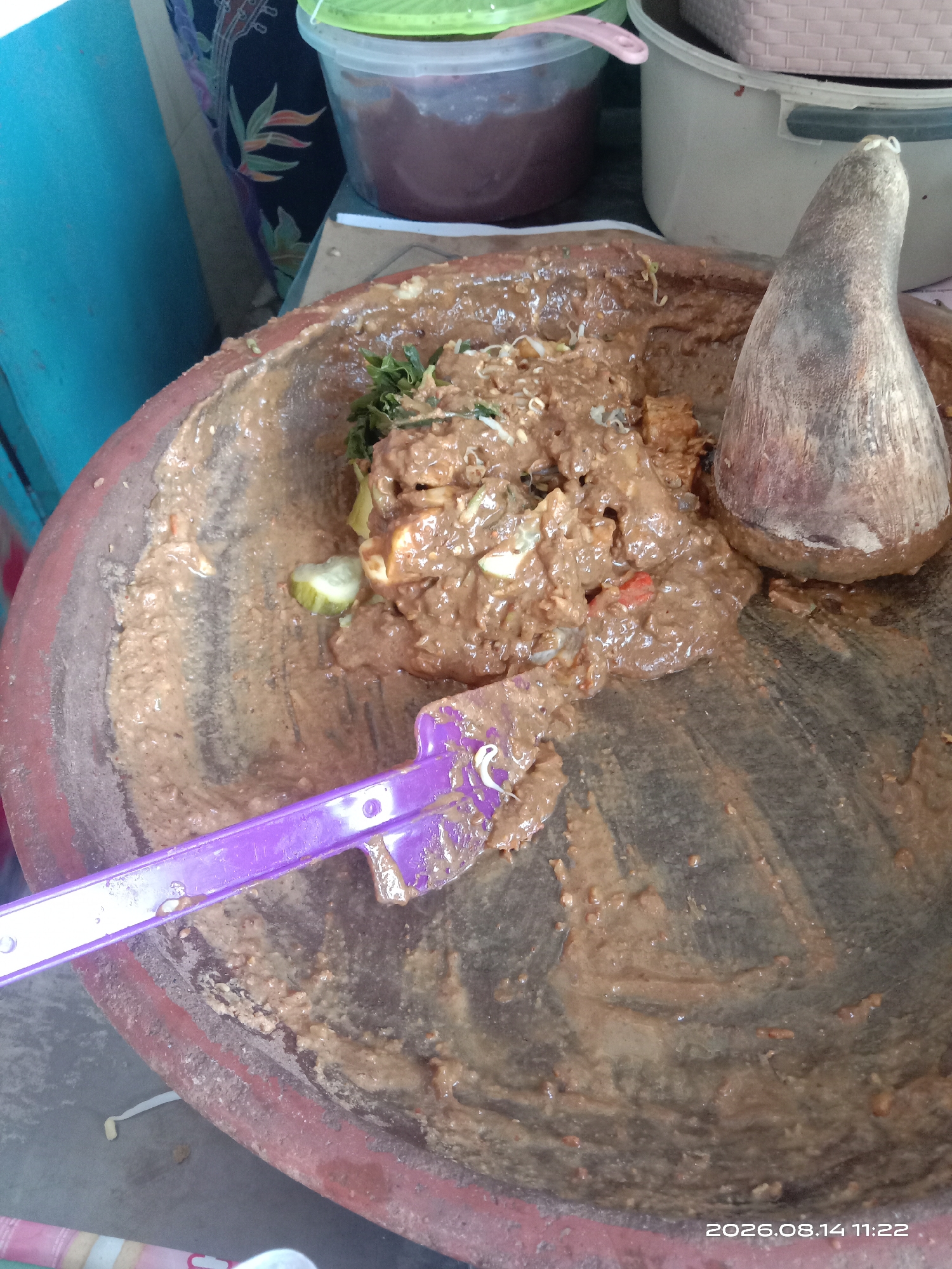 A mortar filled with a brown, peanut-based sauce and vegetables is pictured in a close-up shot. A wooden pestle rests in the mortar, and a purple plastic spatula is partially submerged in the sauce. The mortar appears to be made of stone and is worn from use. In the background, a blue wall is visible, along with a plastic container and a piece of fabric with a floral pattern. A date stamp of "2026.08.14 11:22" is present in the lower right corner of the image. The scene suggests food preparation or serving in what appears to be an outdoor or market setting, possibly in Blimbing, Indonesia, given the context provided. No individuals are visible in the image.