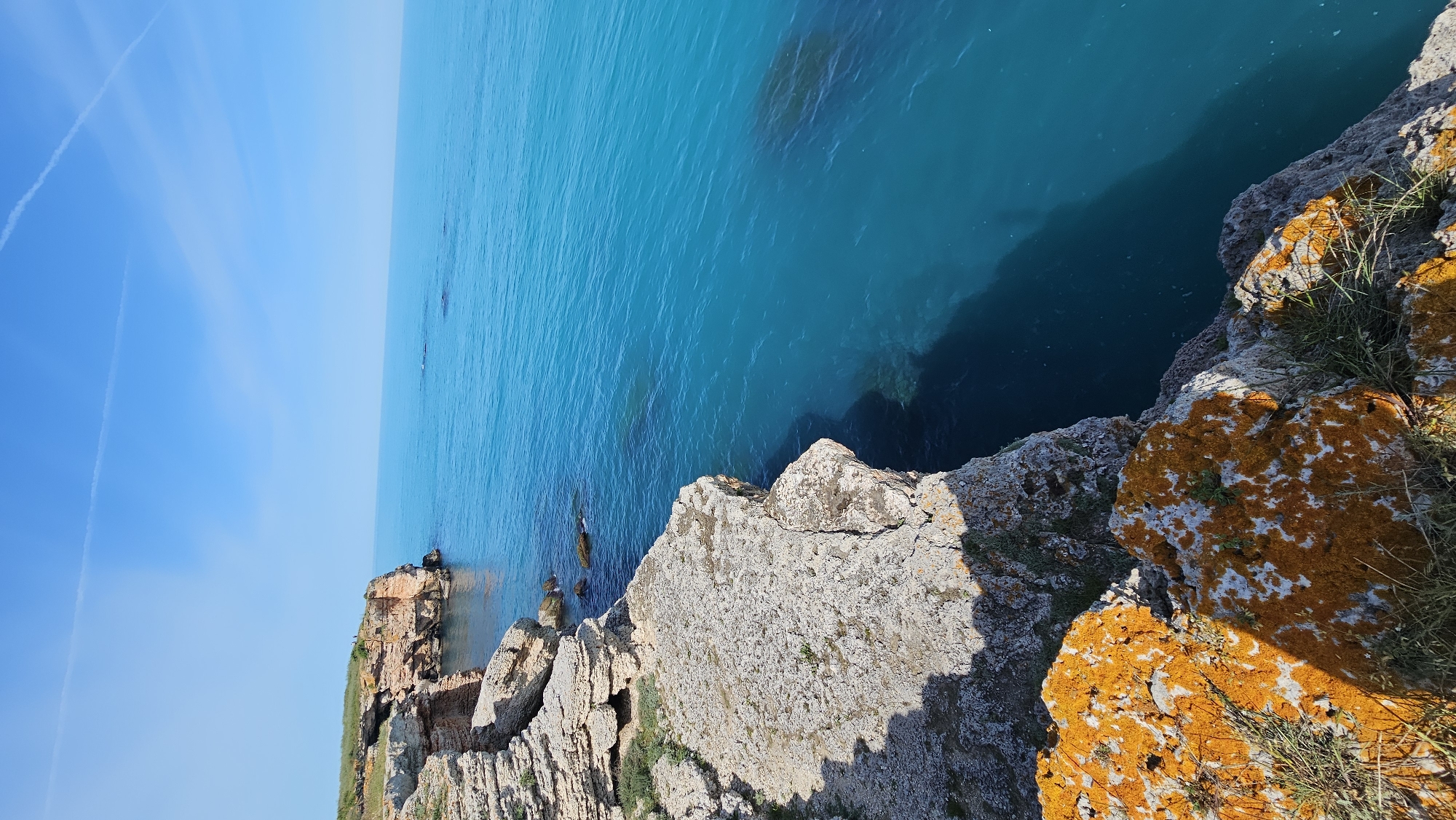 The image captures a scene from Kamen Bryag, Bulgaria. The viewpoint is from a cliff overlooking the Black Sea. The water is a vibrant turquoise, contrasting with the rocky coastline. The cliffs are composed of layered rock formations, some covered with orange lichen and sparse vegetation. The sky is clear blue. In the distance, along the coast, there are more cliff formations and a built structure that could be part of an old fort or staircase. There is no visible human presence in the scene.
