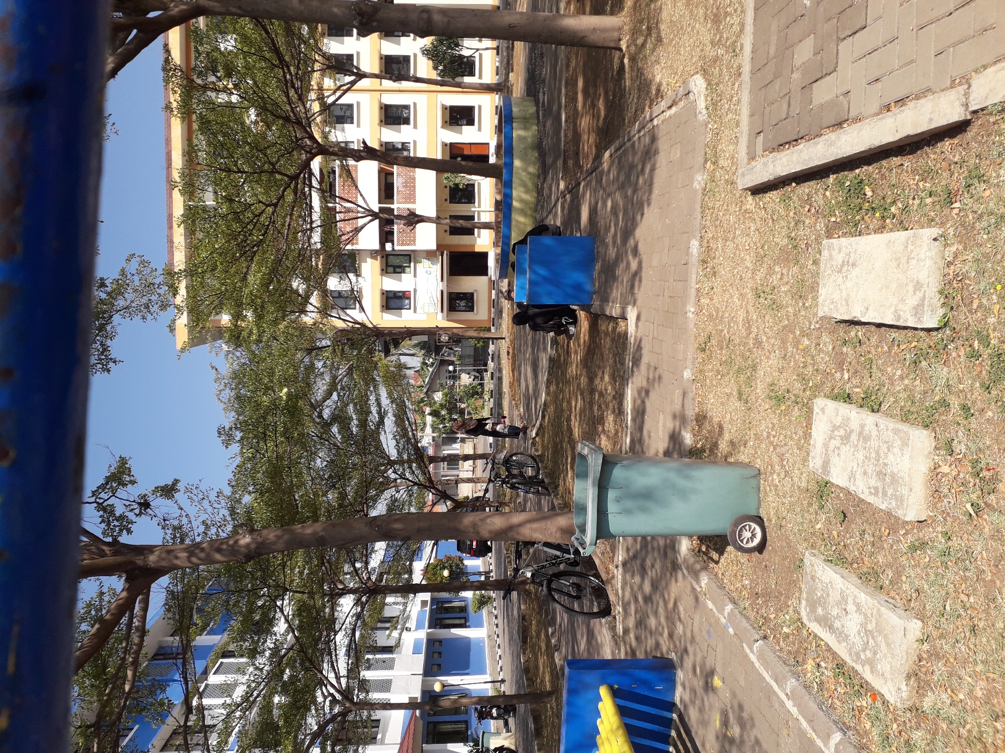 The image depicts an outdoor scene in Cankuang, Indonesia, likely within a residential or campus area. In the foreground, there is a light blue trash bin with a detached wheel lying beside it. Two rectangular concrete slabs are situated on a patch of dry grass and fallen leaves. To the left, a section of blue and yellow playground equipment is visible. Behind this, a paved pathway leads to a light-colored building with blue accents. Several trees with sparse green foliage are scattered throughout the scene, casting shadows on the ground. In the middle ground, two bicycles are parked near the trees. Further back, two multi-story buildings with cream-colored exteriors and orange roofs are visible, with trees in between them. A few individuals are present: one woman in a headscarf is walking near the bicycles, and two people in dark clothing are seated near a blue rectangular structure in the right-middle ground. The sky is clear and blue.