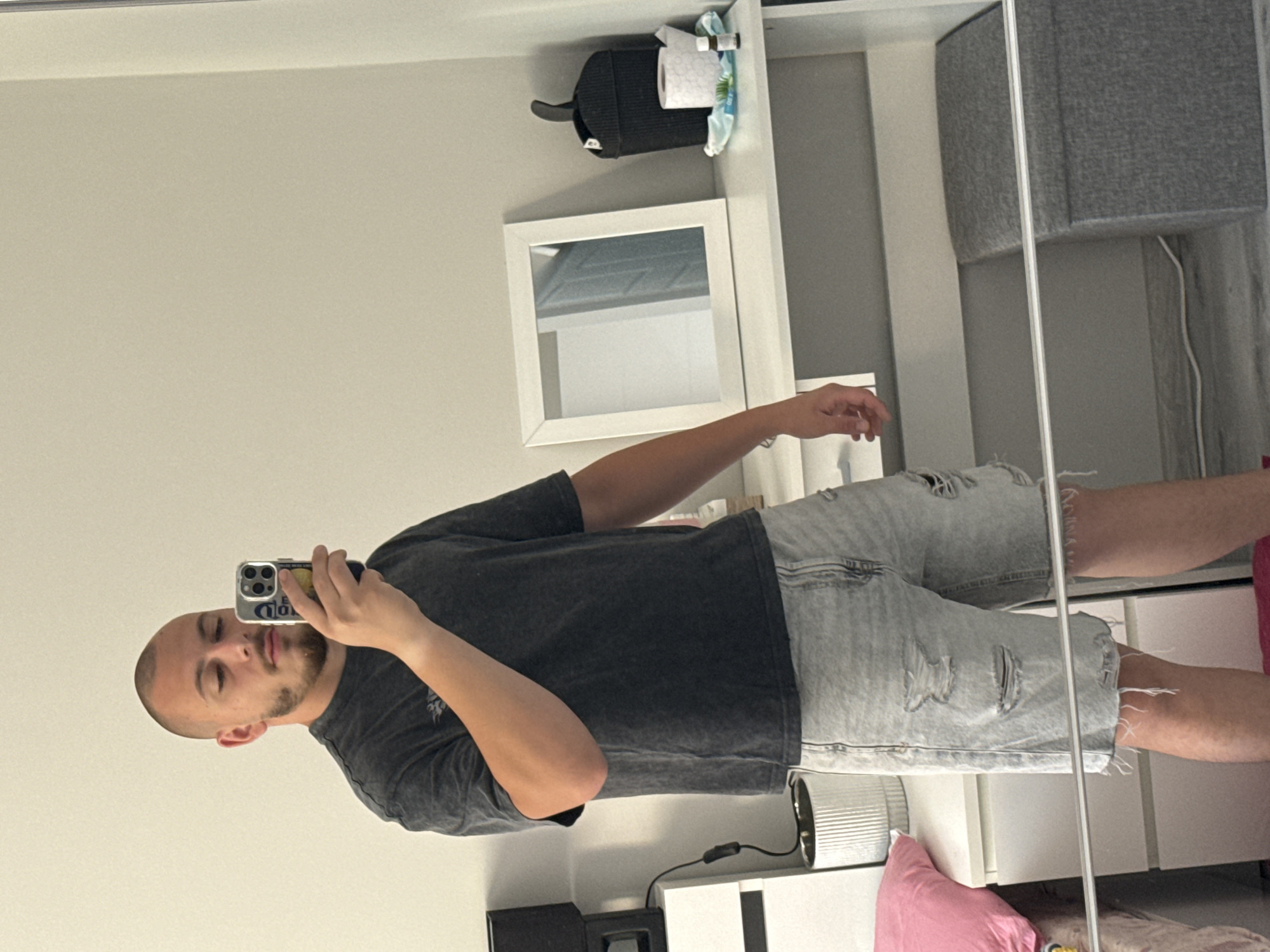 A young man with a shaved head and beard is taking a mirror selfie in what appears to be a bedroom. He is wearing a dark grey t-shirt and light blue distressed denim shorts. He is holding up a smartphone with a case that has multiple cameras.
Behind him, there is a white dresser with drawers, a black lamp, and a white plant pot. To his right, there is a white desk with a square mirror, a black trash can with a roll of toilet paper and a small bottle on top. Part of a grey storage ottoman is visible in the foreground, reflected in the mirror. The walls are a neutral, light color. The floor appears to be grey. There is no indication of the location being in Stara Zagora, Bulgaria.