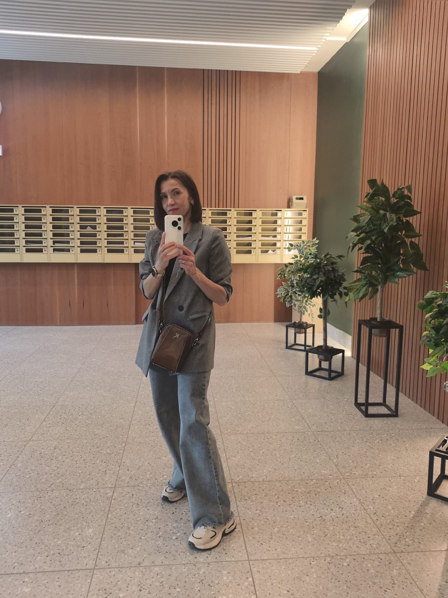 A woman is captured in a selfie, standing in what appears to be a modern building's lobby or hallway. She holds a white smartphone with both hands, angled to photograph herself. She wears a grey, double-breasted blazer over a dark top, light blue, wide-leg jeans, and light-colored sneakers with prominent soles. A small brown crossbody bag with a visible "K" initial on the front hangs over her left shoulder.
The background features walls clad in vertical wooden slats, providing a warm, contemporary aesthetic. Below the wooden paneling, a long series of light-colored mailboxes with small, indistinct labels are arranged in rows. To the right, a section of the wall is painted a muted green, complementing the wood tones. Several artificial potted plants, varying in size, are displayed in black, geometric metal stands, adding decorative greenery to the space. The ceiling features modern strip lighting integrated into slatted panels. The floor is composed of light-colored terrazzo tiles. The overall lighting suggests an indoor setting, likely during the day, illuminated by artificial light sources. The woman's expression is neutral as she looks towards the phone.
