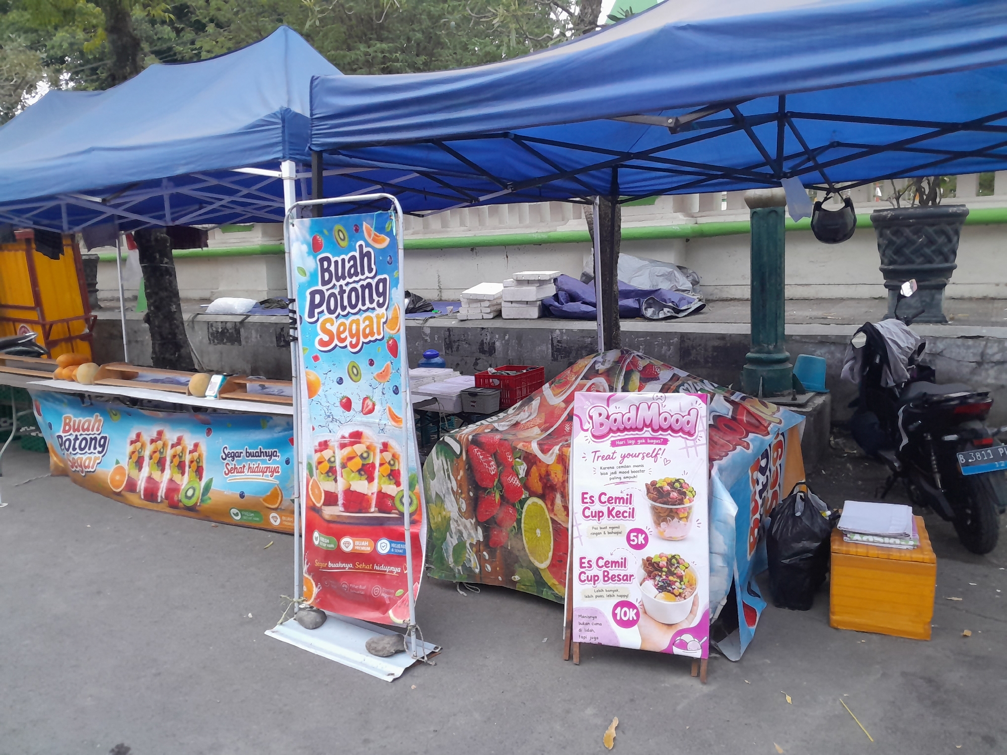 The image depicts a street scene in Slawi, Indonesia, featuring a food stall under a blue canopy. The stall appears to be selling fruit and snacks. Two prominent signs are visible: one displaying "Buah Potong Segar" (Fresh Cut Fruit) with images of fruit cups, and another advertising "BadMood" with various dessert options like "Es Cemil Cup Kecil" (Small Snack Cup) and "Es Cemil Cup Besar" (Large Snack Cup). Several plastic cups filled with colorful fruit salad are arranged on display. Behind the fruit stall, there are stacks of white styrofoam containers and some blue tarpaulin. In the background, a motorcycle is parked, and a large potted plant is visible. The ground is paved with concrete. The overall environment suggests an outdoor market or roadside stall.
