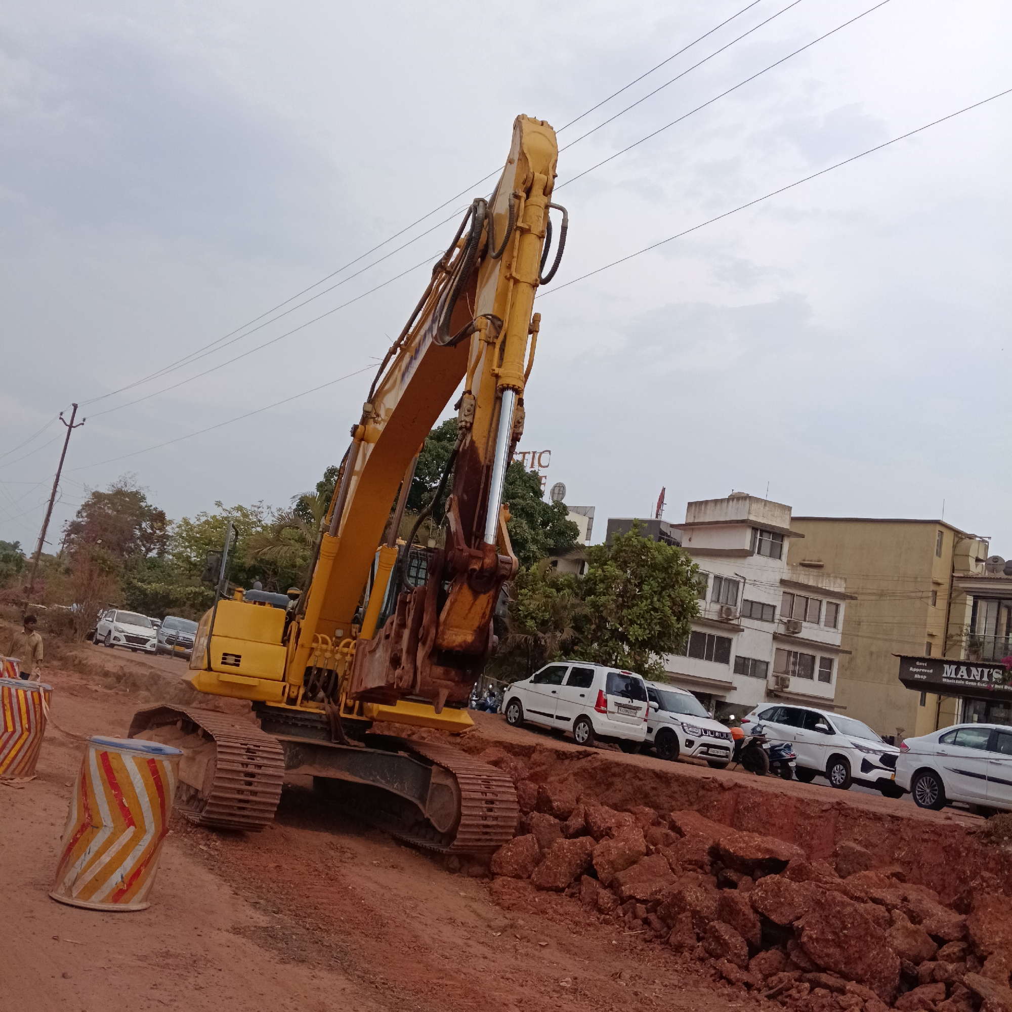The image shows a yellow excavator parked on a dirt road in Britona, India. The road appears to be under construction, with piles of red rocks and debris. A worker is standing near striped traffic barrels to the left of the excavator. Several white cars are parked on a higher road level adjacent to the construction. Buildings and trees are visible in the background. The sky is overcast. Power lines are visible overhead. A sign for "MANI'S Wholesale Cash & Carry" is partially visible on a building in the background.