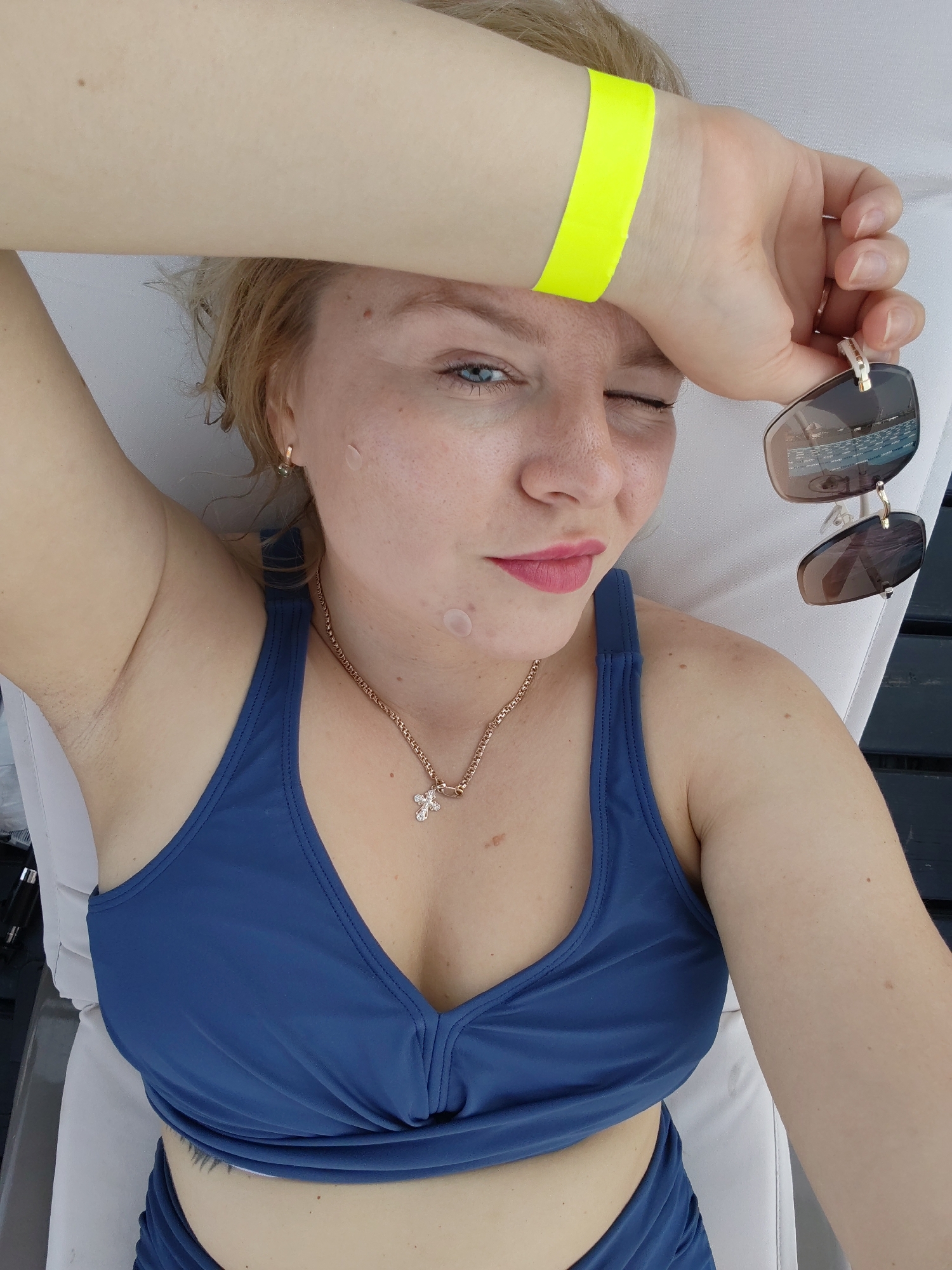 A woman with blonde hair and blue eyes is lying on a white lounge chair. She is wearing a blue swimsuit and a gold necklace with a cross pendant. She is holding a pair of sunglasses in her right hand and has a yellow wristband on her left wrist. Her arm is raised, resting on her forehead. She is winking, and her lips are slightly parted with pink lipstick. There are several small circular bandages on her cheek. The scene appears to be outdoors, possibly at a resort or by a body of water, given the reflection of a pool in her sunglasses. The lighting suggests it is daytime. There is no visible text in the image.