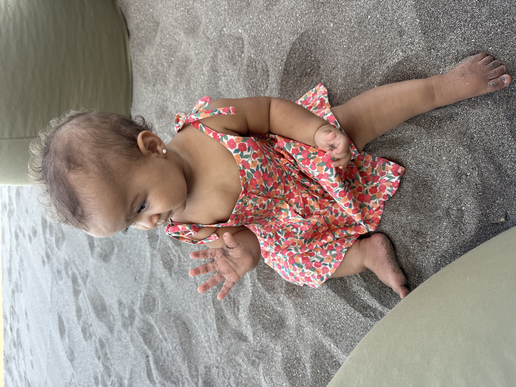 An overhead, slightly angled shot captures a baby sitting on dark grey sand, likely at a beach in North Kuta, Indonesia. The baby, with dark curly hair and a darker skin tone, is dressed in a vibrant, multi-colored floral dress featuring red, orange, and green flowers on a light background, with tie-up shoulder straps. Small white stud earrings are visible in their pierced ears.
The baby is positioned with legs extended and arms bent, looking downwards towards their hands, which are lightly covered in the fine, dark sand. Their left hand is open, fingers spread, showing the sand clinging to their palm and fingertips, indicating interaction with the surface. The sand also dusts their feet.
In the background, on both the left and right sides of the frame, parts of a light green or khaki-colored soft surface, possibly a cushion or outdoor furniture, are visible. The scene is brightly and evenly lit, suggesting daytime with clear weather. The baby appears curious and engaged, exploring the sandy environment. No visible text is present in the image.