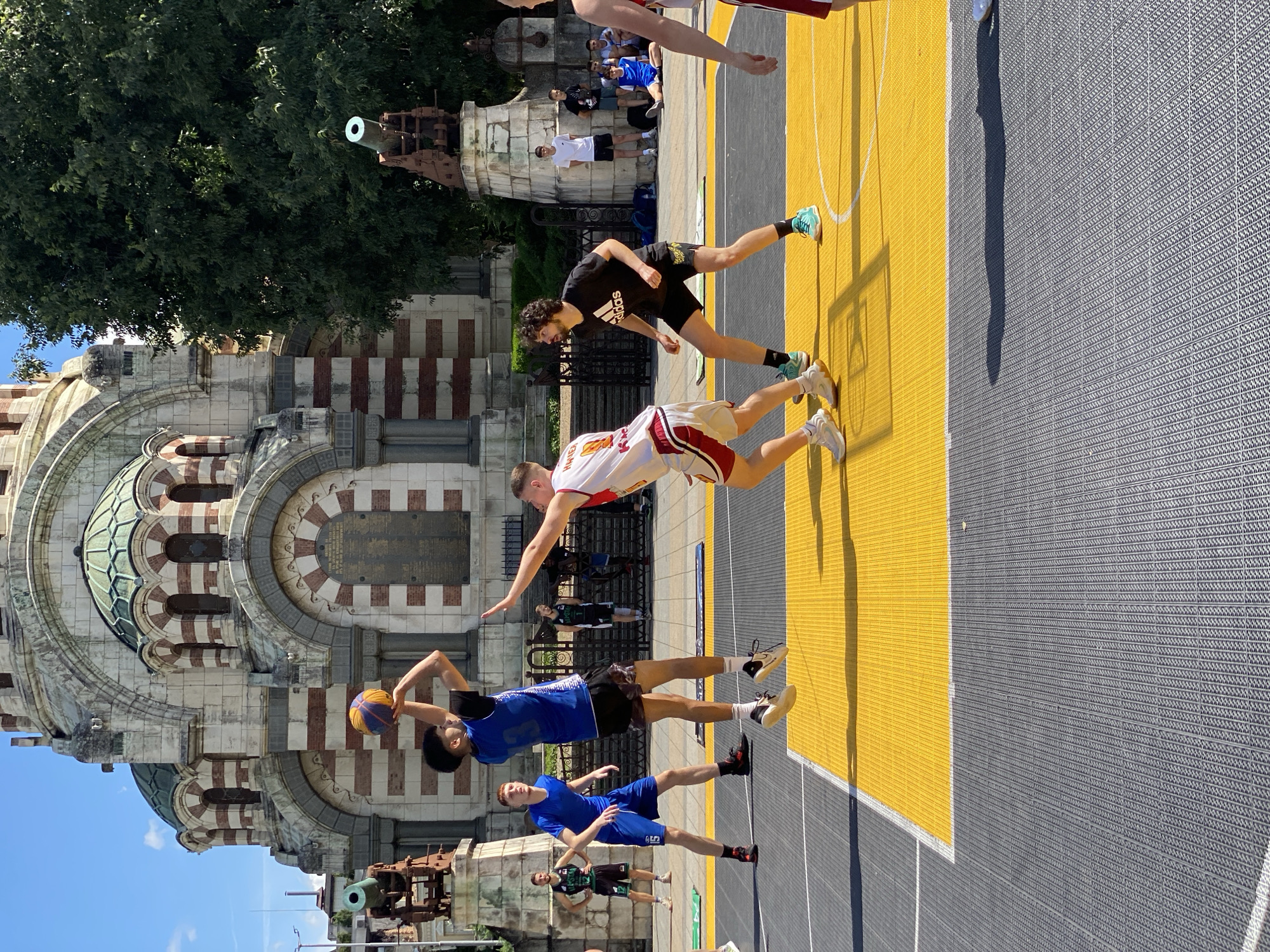 The image depicts a street basketball game taking place on a court in front of the St. Nicholas Military Cathedral in Pleven, Bulgaria. The cathedral, a prominent landmark, features distinctive striped domes and a facade of stone and red brick. Two cannons are visible on pedestals on either side of the cathedral's entrance.
In the foreground, three young men are actively engaged in the basketball game. One player, wearing a blue jersey and black shorts, is in mid-air, preparing to shoot a multicolored basketball. Another player, in a white and red jersey, is guarding him, with his arm extended. A third player, in a black Adidas t-shirt and black shorts, is positioned nearby, observing the play. Other individuals, presumably spectators or teammates, are visible in the background, some sitting on benches and others standing. The court itself is a bright yellow surface bordered by gray tiling. The overall scene is set outdoors under a clear blue sky on a sunny day.