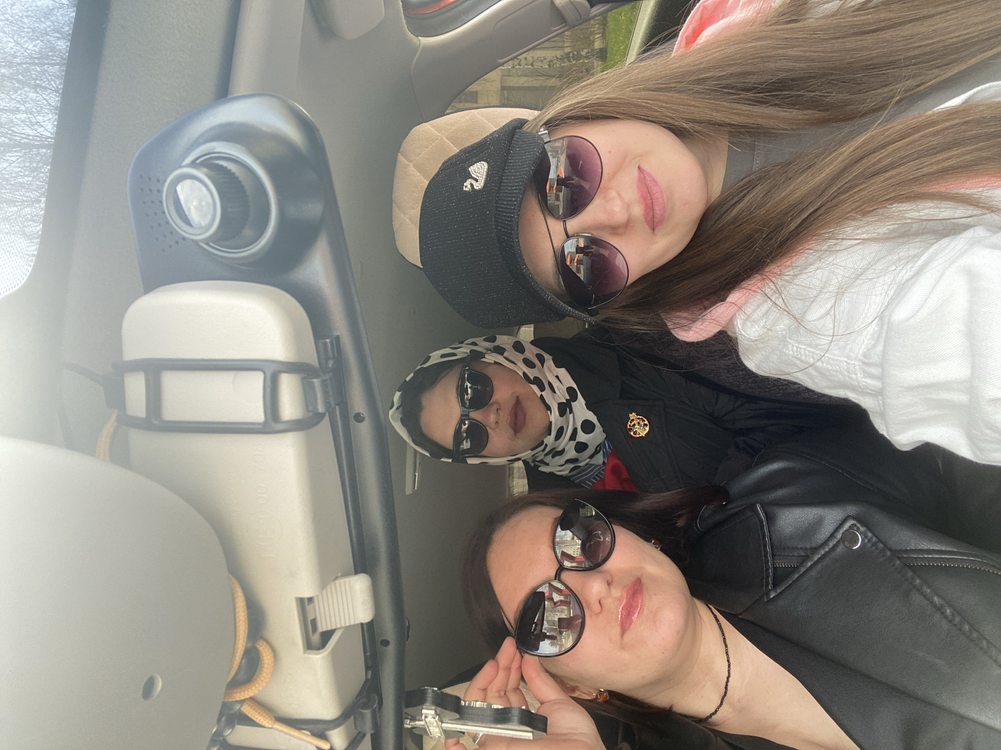 The image shows three women wearing sunglasses, possibly in a car. One woman is wearing a black hat with a swan emblem. Another wears a scarf with a polka dot pattern, while the third is wearing a leather jacket and holding an object in her hand. The environment is a car interior, with a control panel and headrest visible. The location is Марковский, Russia, as the reflections in the sunglasses show buildings that are likely located in that city.