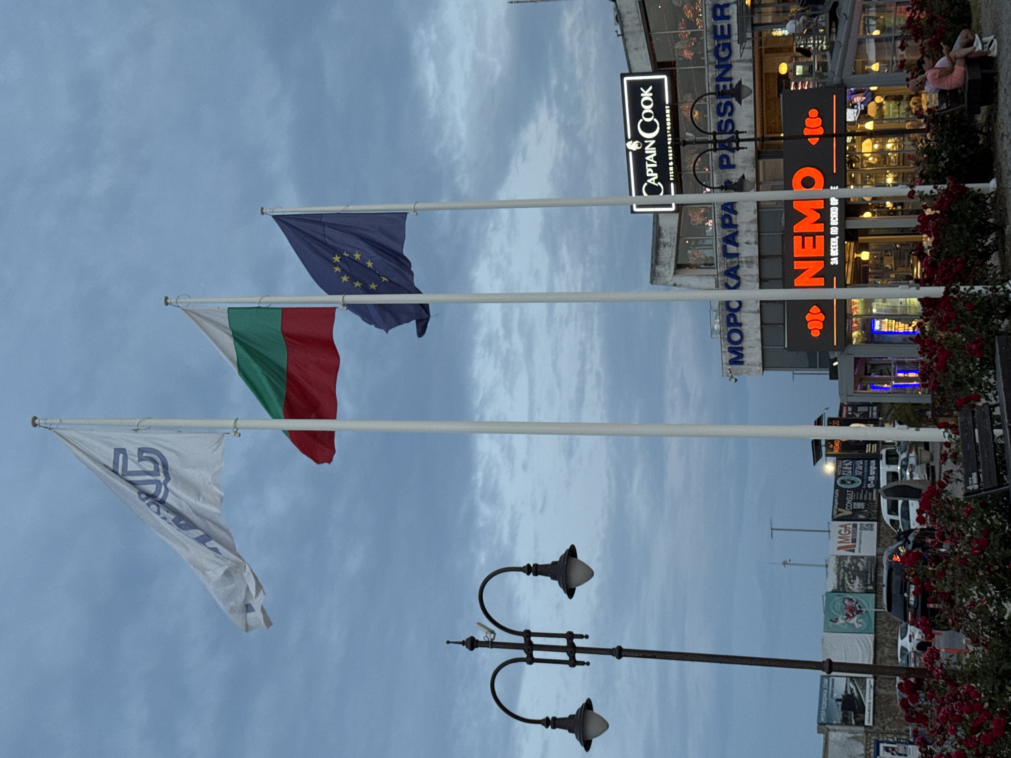 The image depicts a view of a waterfront area in Varna, Bulgaria, likely near the port or a promenade, under a cloudy twilight sky. Three flags are prominently displayed on flagpoles: a white flag with a blue emblem, the Bulgarian flag (white, green, red horizontal stripes), and the European Union flag (blue with yellow stars). In the background, a building complex houses several businesses, including a restaurant named "CAPTAIN COOK FISH & BEEF RESTAURANT" and another establishment with a large neon sign for "NEMO." There are streetlights visible, and a black lamppost with two lights is in the foreground left. In front of the building, there is a paved area with several cars parked. A cluster of red roses is visible in the bottom left corner. Further back, various advertisements and signs are attached to a wall. Two individuals are seated on a bench in the lower right corner, and another person is partially visible in the upper right background. The overall atmosphere suggests an urban waterfront at dusk.