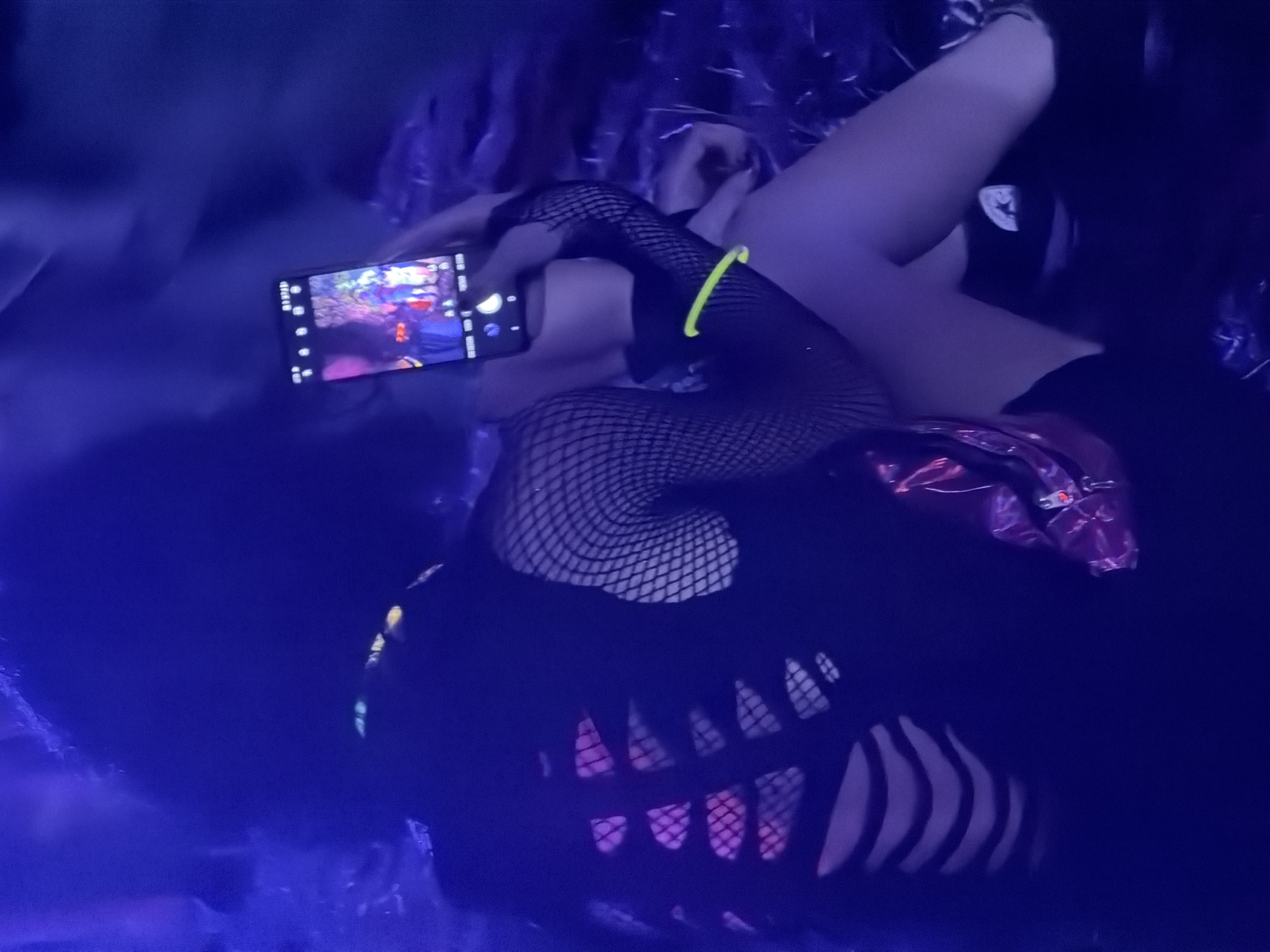 A person is seated in a dimly lit environment, possibly an indoor venue or outdoor night setting with artificial lighting. The person is wearing a black, ripped or cut-out top that exposes parts of their back, revealing pink or purple undergarments or lighting. They are also wearing fishnet stockings and arm sleeves. A metallic pink fanny pack is visible around their waist. The person is holding a smartphone, taking a selfie or recording a video, with the phone's screen illuminated and displaying a colorful image. A neon green glow stick is wrapped around their upper thigh. The background is dark and out of focus, with hints of blue and purple light, creating an atmospheric, possibly party or festival-like setting. The specific location in Echternach, Luxembourg, cannot be determined from the visual information provided.