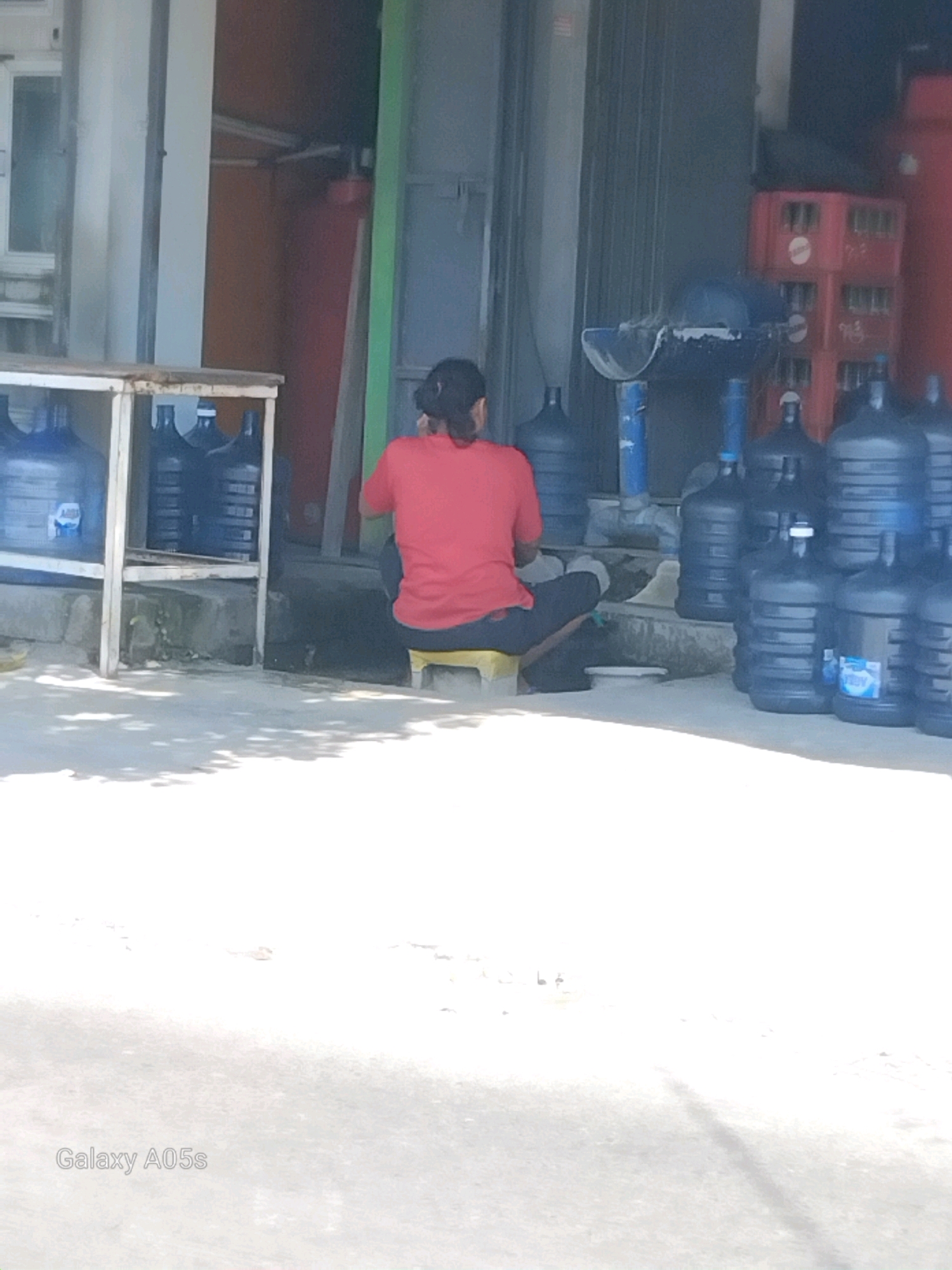 In Karanganyung, Indonesia, a person wearing a red shirt and dark pants sits on a yellow stool outdoors, facing away from the camera. They are positioned in front of a shelf holding several large blue water jugs, with more jugs stacked behind them. To the right, red crates are visible, along with more blue water jugs. The ground is concrete, with dappled sunlight and shadows. A watermark for "Galaxy A05s" appears in the lower left corner.
