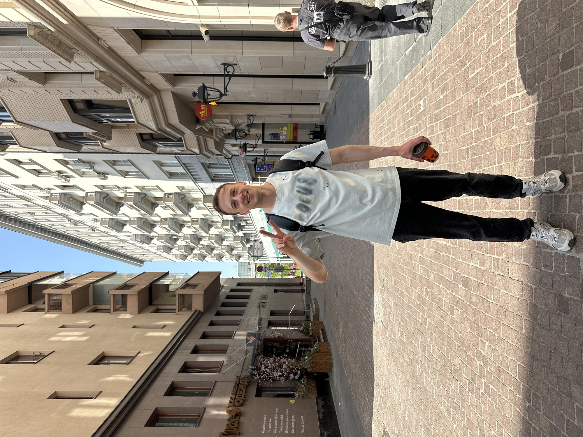 A medium shot captures a young man, smiling at the camera while holding up a peace sign with his left hand and a coffee cup in his right hand. He is dressed in a light-colored t-shirt, dark pants, and light grey athletic shoes. He is also wearing a black backpack. He is standing on a brick-paved sidewalk in a city street. In the background, a blurred figure of a man in a dark uniform can be seen. Buildings line the street, with the building on the left showing a sign in Russian for "Designer Clothing." The text of the poem is visible below this sign. Other storefronts, with some including flowers, are visible. This photograph was taken in Moscow, Russia.
