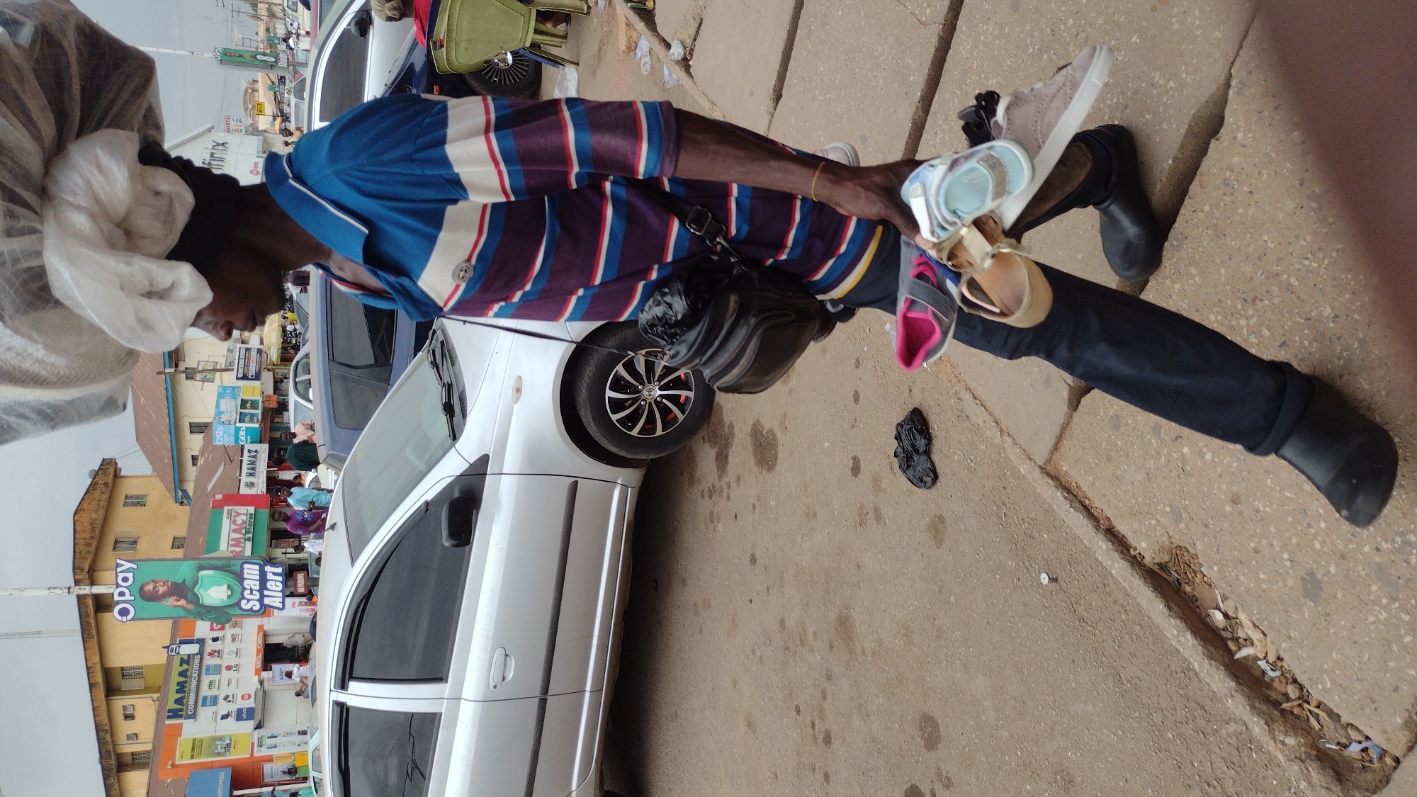 An outdoor scene in Jos, Nigeria, shows a dark-skinned man carrying a large sack on his head. He is wearing a blue and red striped shirt, dark pants, and black shoes. A black bag hangs from his shoulder. He is holding several pairs of children’s sandals and shoes in his hand. A white Toyota car is parked nearby. Debris is on the ground, and a city street with commercial signage is in the background. "Scam Alert" is on a sign. Buildings are visible in the distance.
