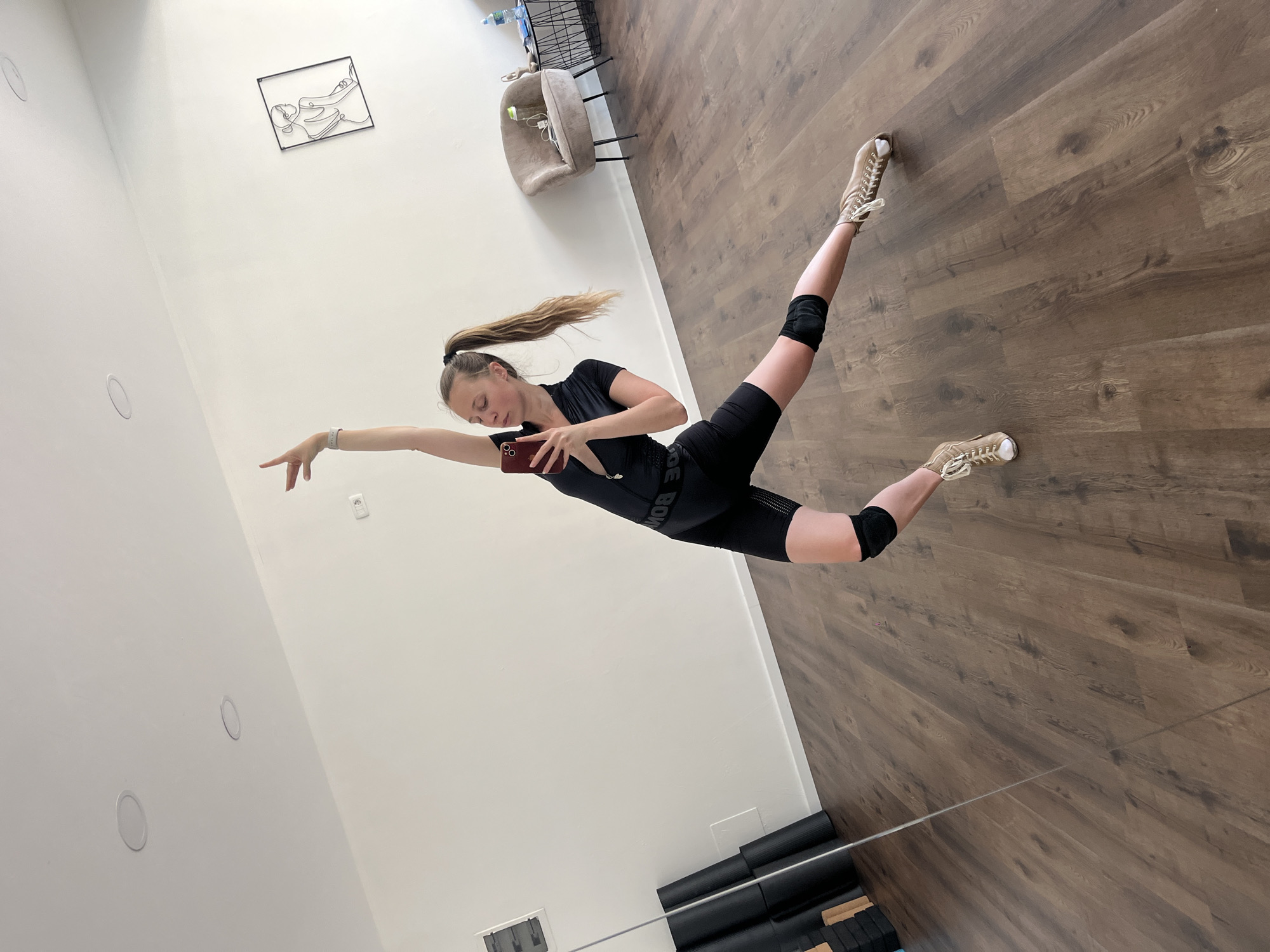 A young woman is captured in a dynamic pose within a brightly lit indoor space, rotated 90 degrees clockwise to create the illusion of her flying horizontally across the room. She has light skin, and her light brown or blonde hair is tied in a high ponytail, which appears to be in motion. She is dressed in black athletic wear, including a short-sleeved top, cycling shorts with the visible text "BOND", and black knee pads. On her feet are beige sneakers with white laces.
Her body is stretched out, with her right arm extended forward, fingers pointed, and her right leg extended backward and slightly upward. Her left arm is bent, holding a dark-colored smartphone with a red case, and she appears to be looking down at its screen. A light-colored watch or fitness tracker is on her right wrist.
The setting features white walls and a dark brown wooden floor. On one wall, there is a framed abstract metal line art piece depicting a reclining figure. In the background, an armchair and a black wire basket containing items like a water bottle are visible. The lower section of the image shows a reflection in a large mirror, revealing a stack of black exercise mats and possibly some yoga blocks. Two circular recessed lights are visible on the white wall, and a small white remote control is also present. The bright, even lighting suggests it is daytime. The overall ambiance points to a studio or gym environment.