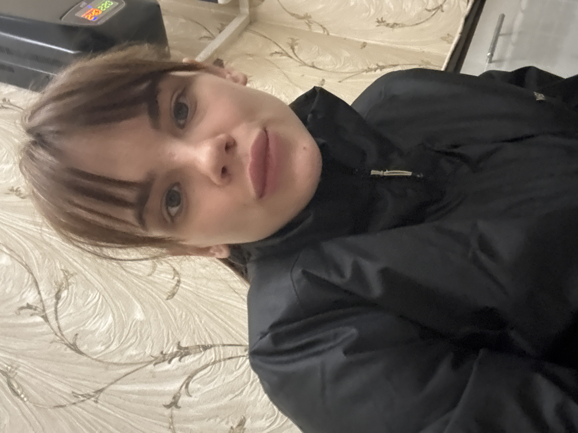 A selfie shows a person with short brown hair and bangs wearing a black jacket. They are facing the camera with a slightly angled pose. The background appears to be a wall with a patterned wallpaper, likely indoors. In the upper left corner, there is a device with a digital display showing numbers "210220". The location is unknown and cannot be deduced from the image. The city of Rostov-on-Don, Russia, is not discernible.