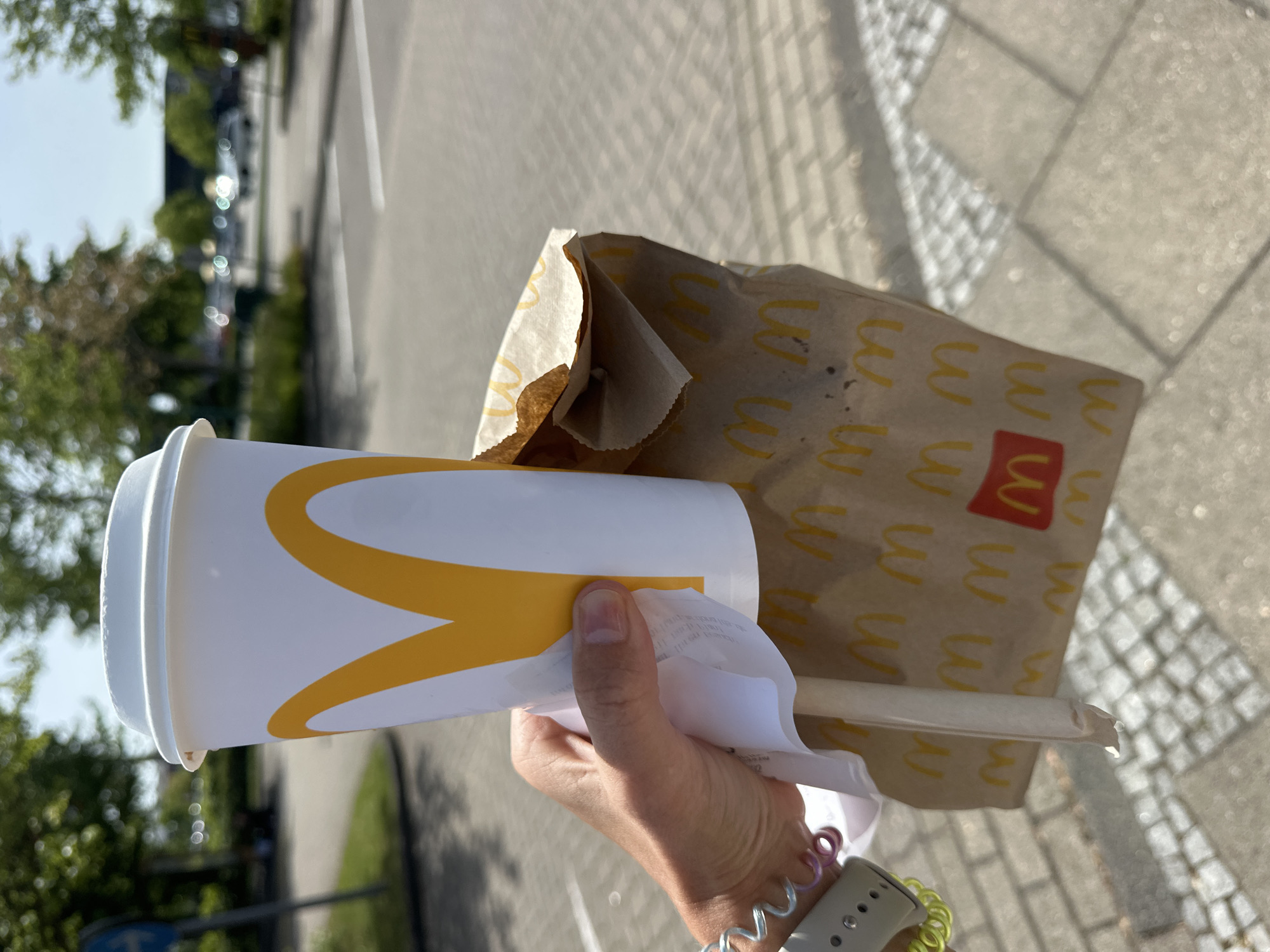 A person holds a McDonald's cup with a golden arches logo and a brown paper bag with multiple golden arches and a red McDonald's logo, likely containing food. A wrapped straw is protruding from the bag. The person is wearing a wristwatch with colorful bands. The background shows a parking area with cars, trees, and a paved walkway. The location is likely in or near Ottersberg, Germany, as inferred from the general context of McDonald's locations.