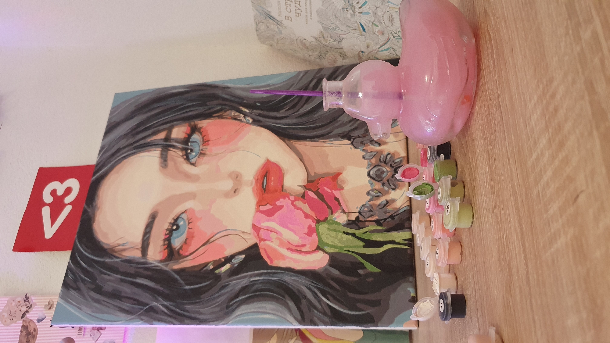 A paint-by-numbers kit featuring a woman's face, a pink water bottle with a straw, and a collection of small paint pots are placed on a wooden surface. The woman in the painting has dark hair, blue eyes, and is holding a pink flower in her mouth. The red object with "<3" on it and the printed book are on the left side of the painting. There is no evidence to suggest that this scene is in Bar, Montenegro, or anywhere else.