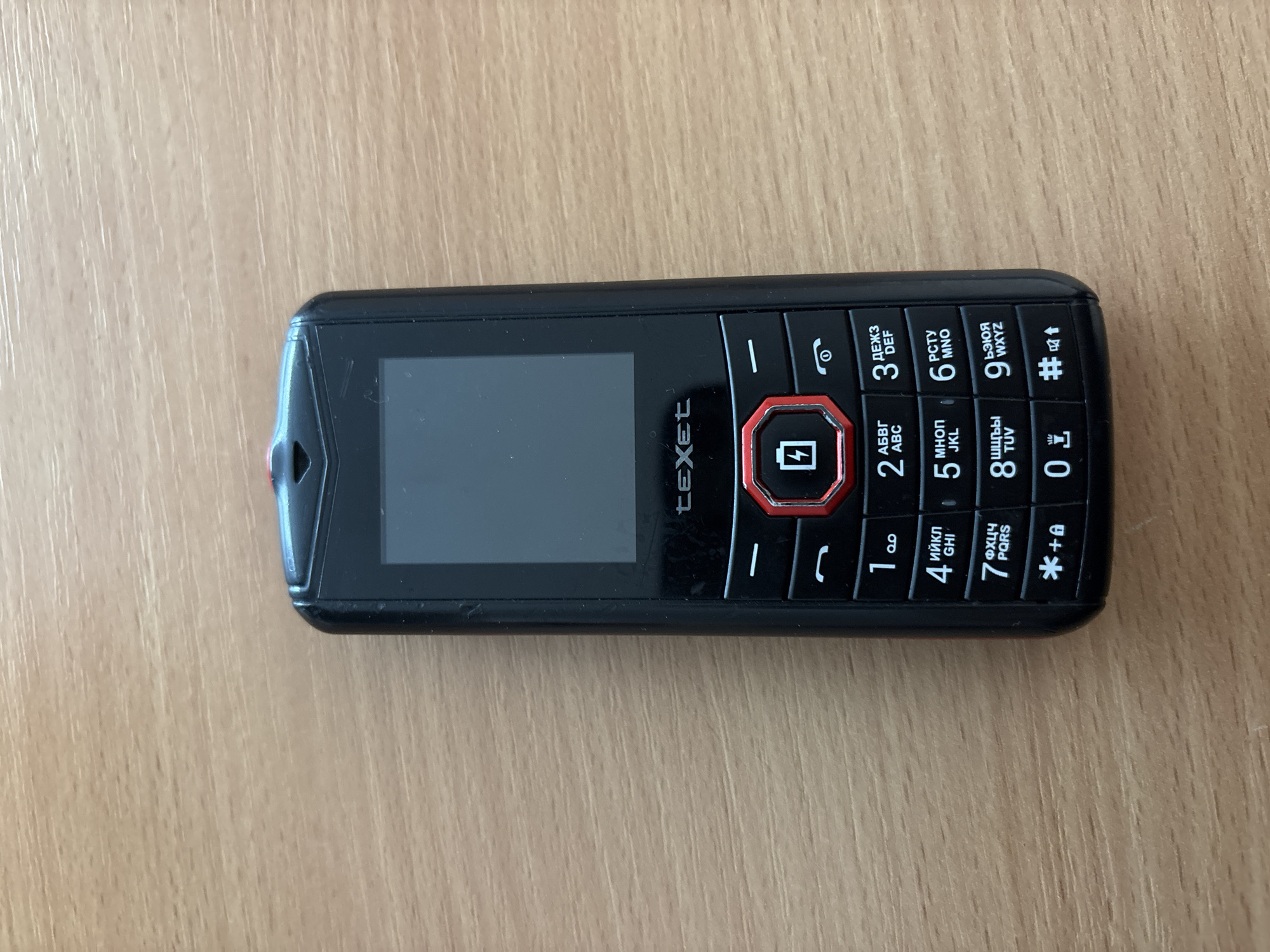 A black Texet feature phone is lying on a wooden surface. The phone has a screen at the top and a physical keypad below. The keypad has numbers 0-9, as well as symbols like asterisk, pound sign, and a power button. The background is a wooden desk. The image does not provide any information about the city of Minsk, Belarus.