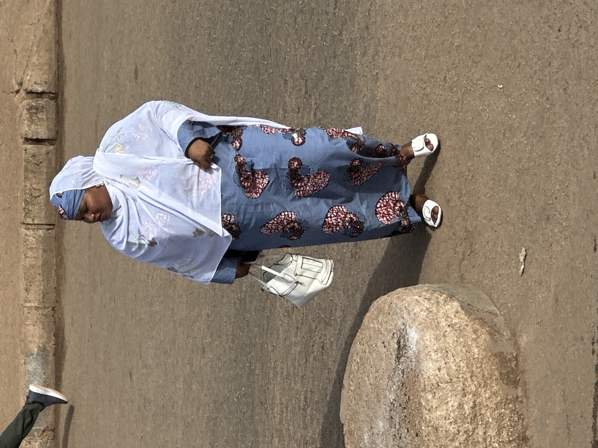 An overhead shot shows a woman in Bukuru, Nigeria, wearing a light blue dress with a floral pattern of dark pink and white. She has a light blue headscarf and a white shawl draped over her shoulders. The woman is carrying a white handbag in her right hand and is wearing white sandals. She is standing on a dirt or gravel surface. To the left of the woman, a large, light brown, rough-textured stone object is visible. In the upper left corner of the image, the lower legs and feet of another person wearing dark pants and black sneakers are visible, suggesting this person is walking. The background appears to be a paved area with some textured blocks or stones.