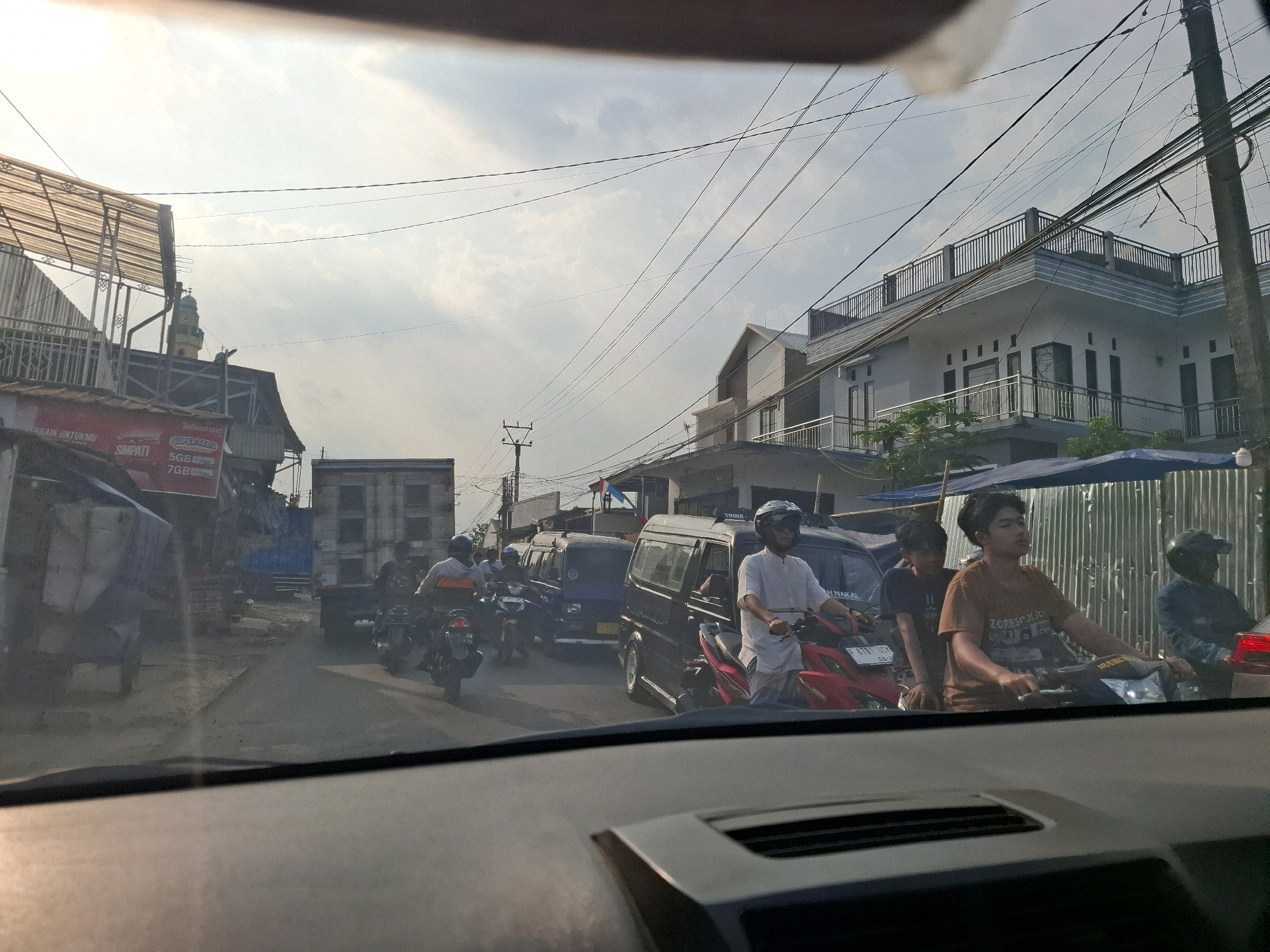 The image captures a street scene in Sukabumi, Indonesia, likely during daytime, as indicated by the bright sky and shadows. The perspective is from inside a vehicle, looking forward through the windshield. The street is populated with various vehicles, including motorcycles, vans, and a truck. Several people are visible, predominantly riding motorcycles.
In the foreground, the dashboard of the vehicle from which the photo is taken is visible, along with the lower part of the windshield. The road surface appears to be asphalt, and there is moderate traffic congestion.
On the left side of the street, there are buildings and structures, including a building with a metal awning and what appears to be a shop or market. A red and white banner with text in Indonesian is also visible. Further down the street, a mosque with a minaret is partially visible.
The right side of the street features modern buildings, one of which is a multi-story white house with balconies and railings. Overhead, a complex network of power lines crisscrosses the sky, supported by utility poles.
Specific details about people include:
- A person wearing a white shirt and helmet is riding a red motorcycle in the center-right of the image.
- Another person wearing a brown t-shirt is on a motorcycle behind the person in the white shirt.
- A person wearing a blue jacket and helmet is on a motorcycle further to the right.
- Several other individuals are on motorcycles in the background, some appearing to be drivers and others passengers.
Objects of note include:
- Various motorcycles, some of which are prominent in the foreground and mid-ground.
- A black van and a blue van are positioned in the traffic.
- A large truck is visible in the mid-ground.
- Utility poles and numerous overhead electrical wires are a dominant feature of the environment.
- Buildings of varying architectural styles, from simple market stalls to modern residences.
The actions observed are primarily related to driving and commuting, with people operating motorcycles and vehicles in traffic. The overall impression is a busy urban street with typical daily activity.