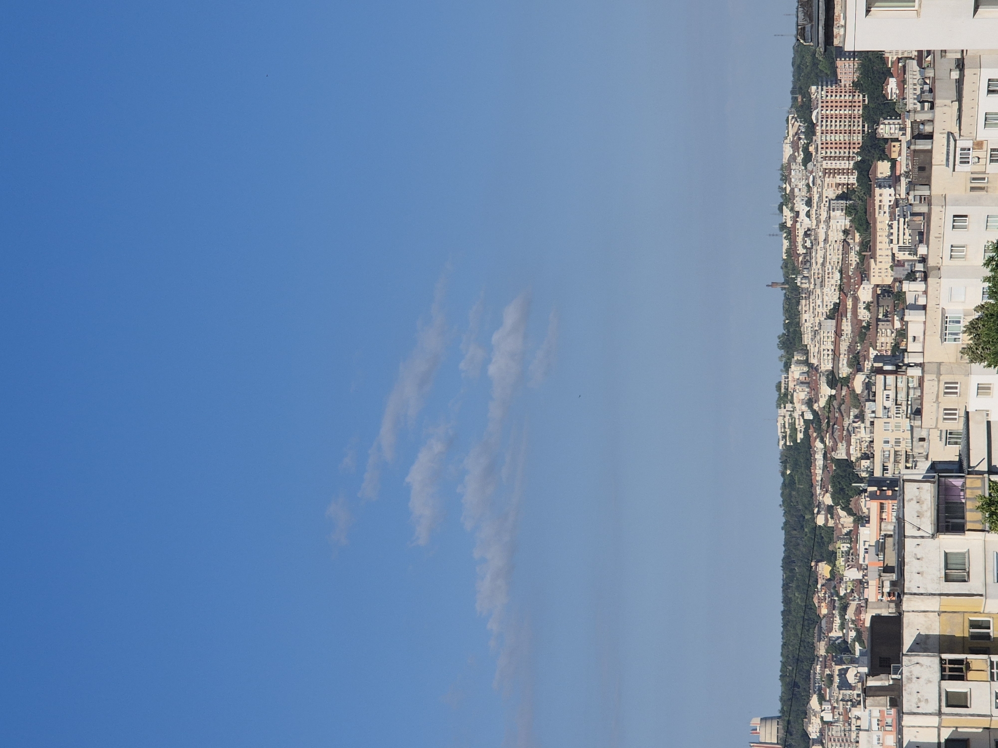 This is a medium-shot, high-angle photograph of a densely populated urban area with a clear blue sky. The city is built on a hillside, with buildings of varying heights and architectural styles. The foreground features several apartment buildings, some with weathered facades, showing signs of age. These buildings are characterized by their simple, rectangular shapes, numerous windows, and balconies.
In the mid-ground, the cityscape extends further up the hill, with a dense collection of residential buildings. Many of these buildings have red-tiled roofs. A notable feature in the mid-ground is a taller, modern-looking apartment building with a grid of windows, contrasting with the older structures.
The background is dominated by a green, tree-covered hill that acts as a natural backdrop to the city. On the very top of the hill, some structures are visible, including what appears to be a tall, cylindrical industrial building or silo on the left, and other smaller buildings scattered amongst the trees. The sky is a vibrant, cloudless blue, with a few faint, wispy clouds in the upper left quadrant. There are no people or animals visible in the frame. The overall impression is one of a sprawling, established city built on uneven terrain.