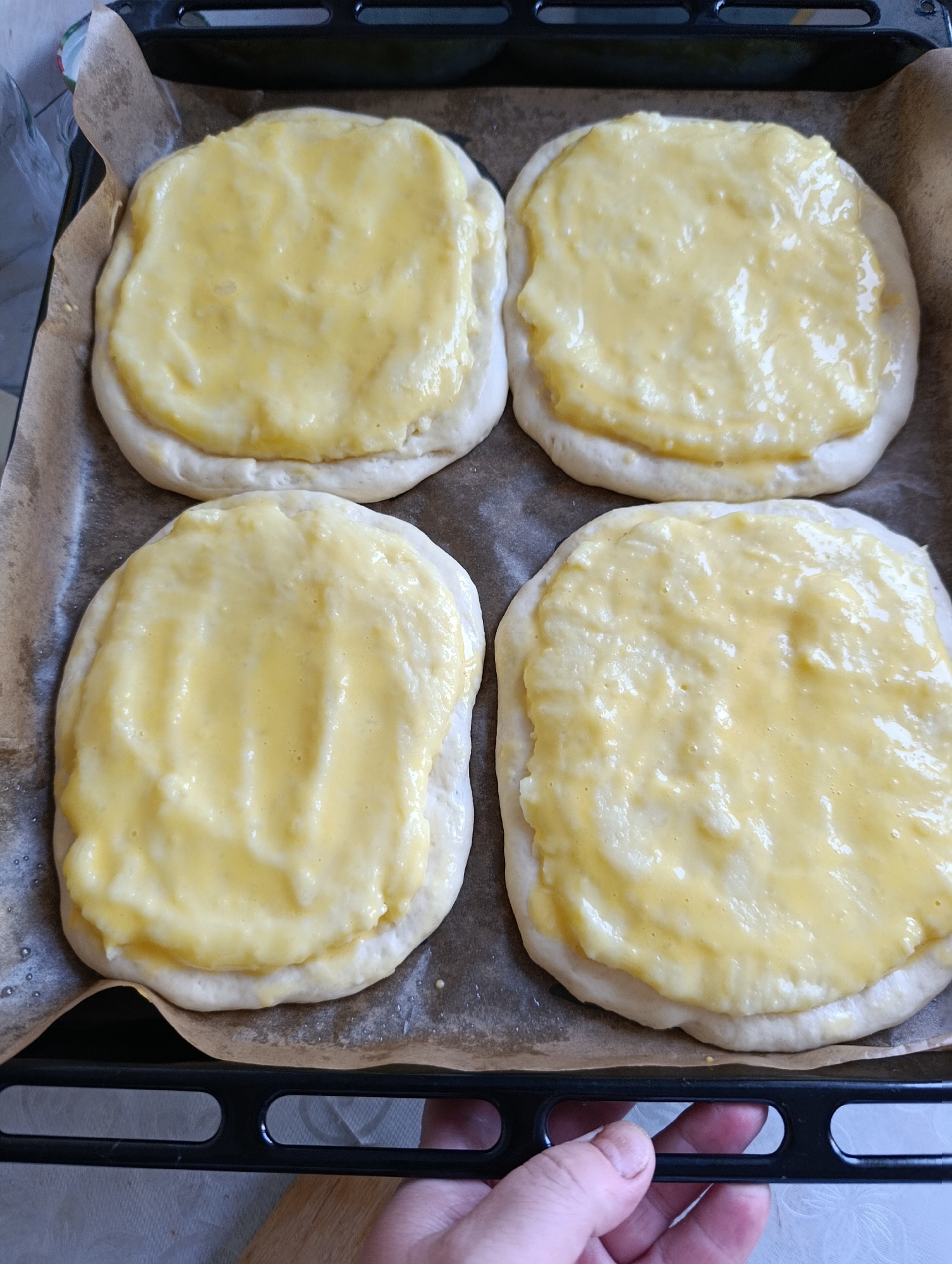 Four round pastries with a light-colored dough base and a creamy yellow topping are arranged on a baking sheet lined with parchment paper. The pastries are placed in a two-by-two grid. The topping appears to be a sweet filling, possibly custard or cheese-based, with a slightly lumpy texture. The baking sheet is black with a textured surface and is placed on a light-colored surface that has a subtle floral pattern. A hand with visible fingernails is holding the edge of the baking sheet from the bottom right. The image appears to be taken indoors. The provided location of Chaikovsky, Russia, is extraneous information to the visual content of the image.