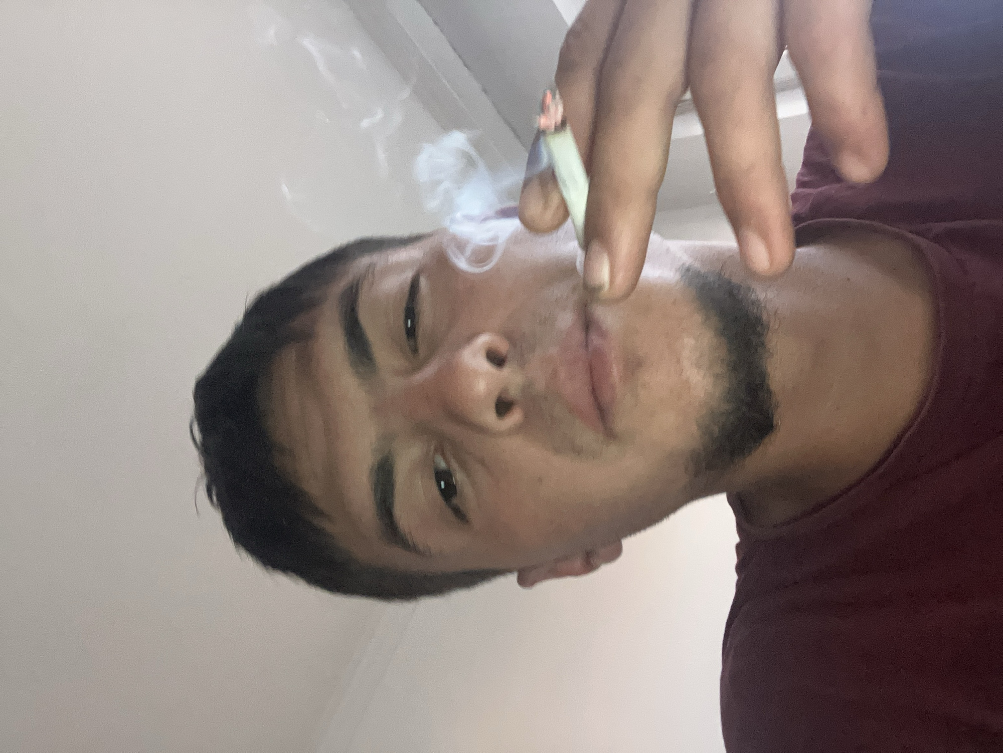 A man is holding a lit cigarette between his fingers, with smoke rising from it. The man has short dark hair and a short dark beard, and is wearing a maroon shirt. The background is a plain white wall. The location of this image is not determinable based on the provided information and image.