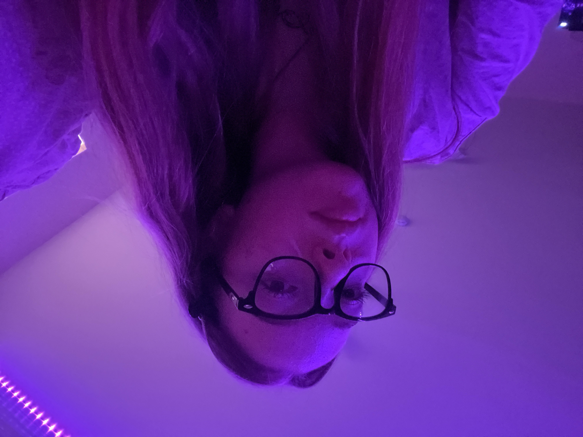 A woman with fair skin and long, reddish-blonde hair is seen upside down, wearing black-framed glasses. She appears to be indoors, illuminated by purple LED strip lights visible in the lower left of the frame. The background is a light-colored surface, likely a wall or ceiling, also washed in the purple light. There are no immediate indicators of the location being Chaikovsky, Russia. The woman is looking down, her face dominating the frame. She is wearing a light-colored long sleeve top. There are no observable actions or interactions.