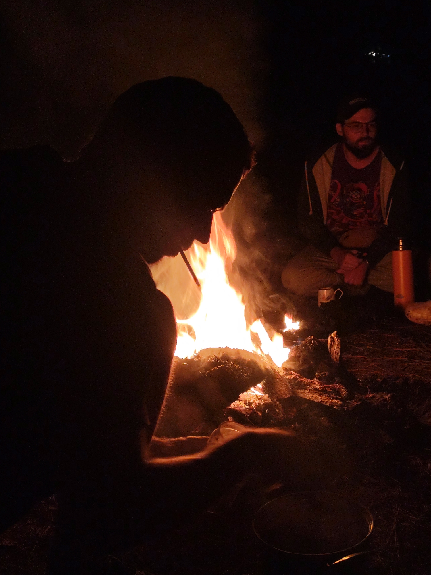 The image depicts a nighttime scene around a vibrant campfire in an outdoor setting in Lạc Dương, Vietnam, with two individuals present. The central element is a brightly burning campfire, characterized by orange and yellow flames and a plume of smoke, serving as the primary light source.
In the foreground, on the left, a person is seen largely in silhouette, their profile sharply outlined by the intense glow of the fire. They appear to be holding a white object, possibly a cigarette or straw, in their mouth. Their right arm is illuminated by the fire, showcasing some hair on the forearm. A dark, circular pot or pan is visible on the ground directly in front of them.
To the right of the campfire, a second person is seated cross-legged on the ground. This individual wears a dark cap, glasses, a beard, and a dark t-shirt featuring a reddish graphic design that appears to be a stylized diving helmet. They are also wearing a lighter-colored hoodie. Their expression is calm and contemplative as they look towards the fire. Adjacent to them on the ground are an orange thermos or bottle and a white mug.
The ground surrounding the fire is composed of dirt and scattered with pieces of wood and dry foliage. The background recedes into darkness, with subtle hints of distant terrain, possibly hills or mountains, visible in the upper left portion of the frame. The overall atmosphere is one of peaceful relaxation, suggesting a camping trip or an evening gathering by the fire in a natural environment.