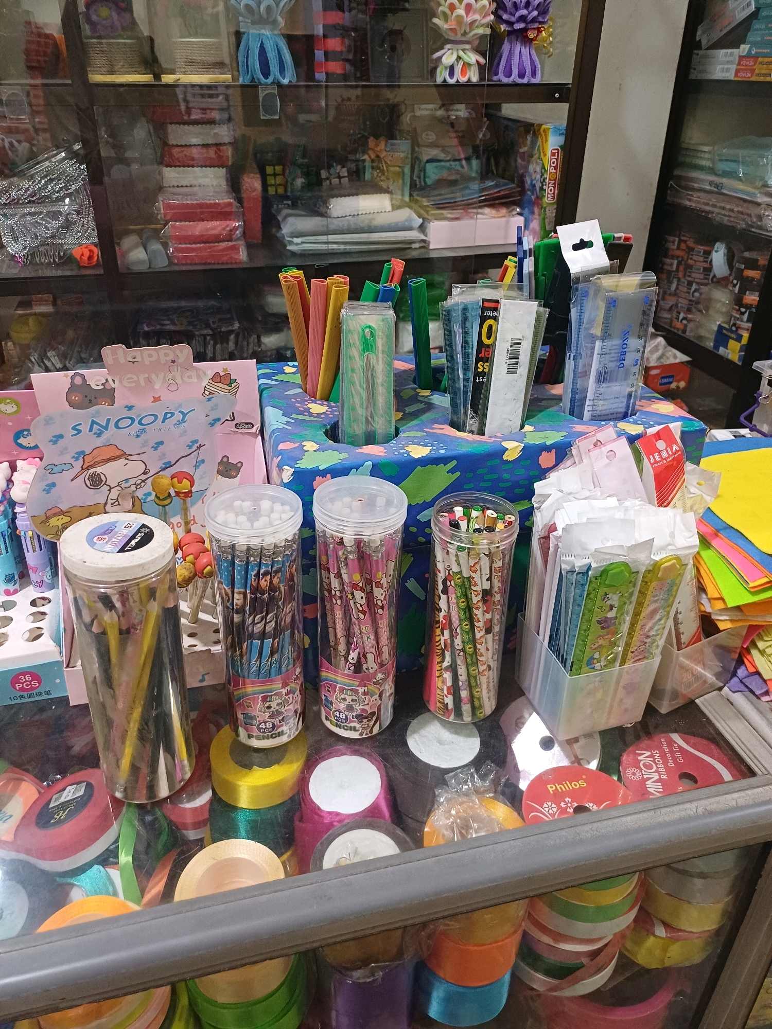 The image depicts a display of art and craft supplies in a shop. The items are arranged on shelves and in containers, suggesting a retail environment. Visible items include pencils, rulers, ribbons, and decorative craft materials. The shop appears to be well-stocked with a variety of colorful items, catering to artistic and crafting needs. The setting is indoors, with glass shelves displaying the merchandise. The lighting suggests an interior artificial light source.