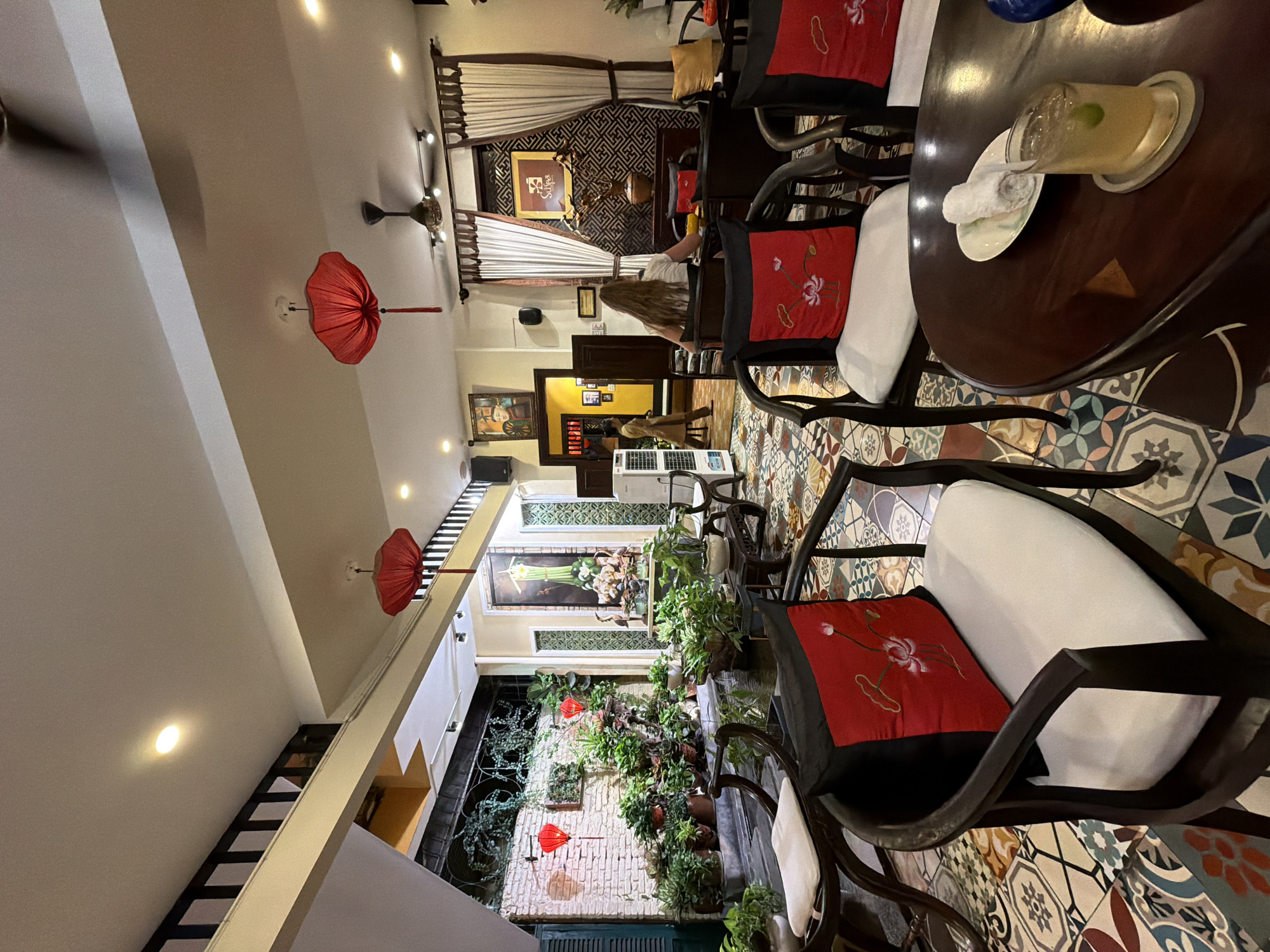 This image depicts the interior of a café in Nha Trang, Vietnam. The café features a distinct Vietnamese aesthetic with traditional red lanterns hanging from the ceiling, a wall adorned with potted plants, and intricately patterned tiles on the floor. Several dark wood chairs with red and black cushions are arranged around dark wood tables. In the background, a woman with long blonde hair is seated at a table, facing away from the camera, while another woman walks through a doorway. The overall atmosphere is one of calm and cultural immersion.