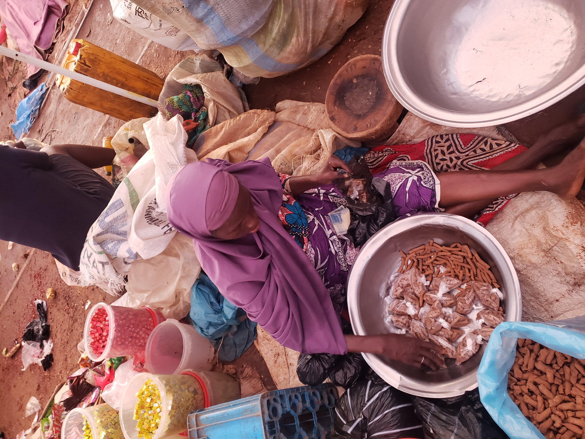 An overhead shot captures a street market scene in Kakuri Gwari, Nigeria. In the foreground, a woman wearing a purple hijab and a patterned dress is seated amidst various goods. She is sorting through small, individually bagged, brown food items placed in a large silver bowl. To the left of the bowl, a blue plastic bag is filled with similar brown food items. Surrounding the woman are several large sacks, black plastic bags, and smaller containers filled with other market goods, including what appears to be red berries or seeds in a clear plastic bucket. A rustic wooden chopping board rests nearby. The ground is covered with dirt and scattered debris, indicative of an outdoor market. In the background, a yellow plastic jerrycan and a blue plastic crate are visible. The overall impression is one of bustling commerce in a typical Nigerian street market.