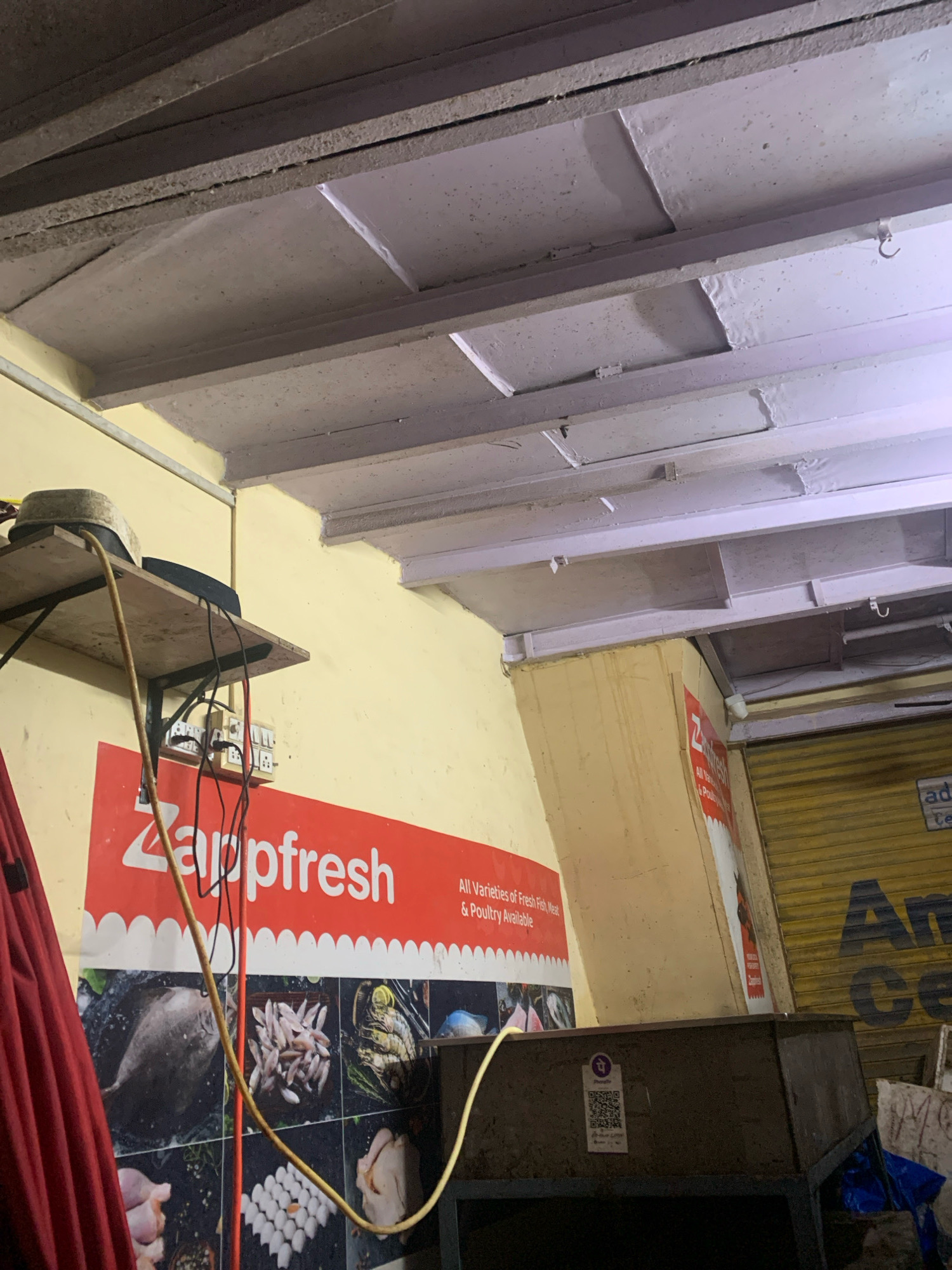 The image shows an interior scene, likely a shop or market stall, in Mumbai, India. The foreground features a large red sign with white text that reads "Zappfresh" and "All Varieties of Fresh Fish, Meat & Poultry Available." Below this text, a collage of images displays various types of fresh food, including fish, eggs, and poultry. A red curtain is partially visible on the left side of the image.
In the mid-ground, a wooden shelf is mounted on a yellow wall. On the shelf, there are two objects, one appearing to be a hat and another dark, possibly electronic device. Electrical wires and a power strip with multiple outlets are attached to the wall below the shelf. A yellow electrical cable snakes across the image, connecting to something out of frame.
The background reveals a yellow-painted wall that curves into a corner. To the right, a closed yellow roller shutter door is visible, with partially obscured blue lettering on it that seems to say "Am" followed by other characters. The ceiling, which is visible in the upper portion of the image, appears to be constructed of a wooden frame with panels, painted in a light purple hue. The overall impression is that of a commercial establishment specializing in fresh food.